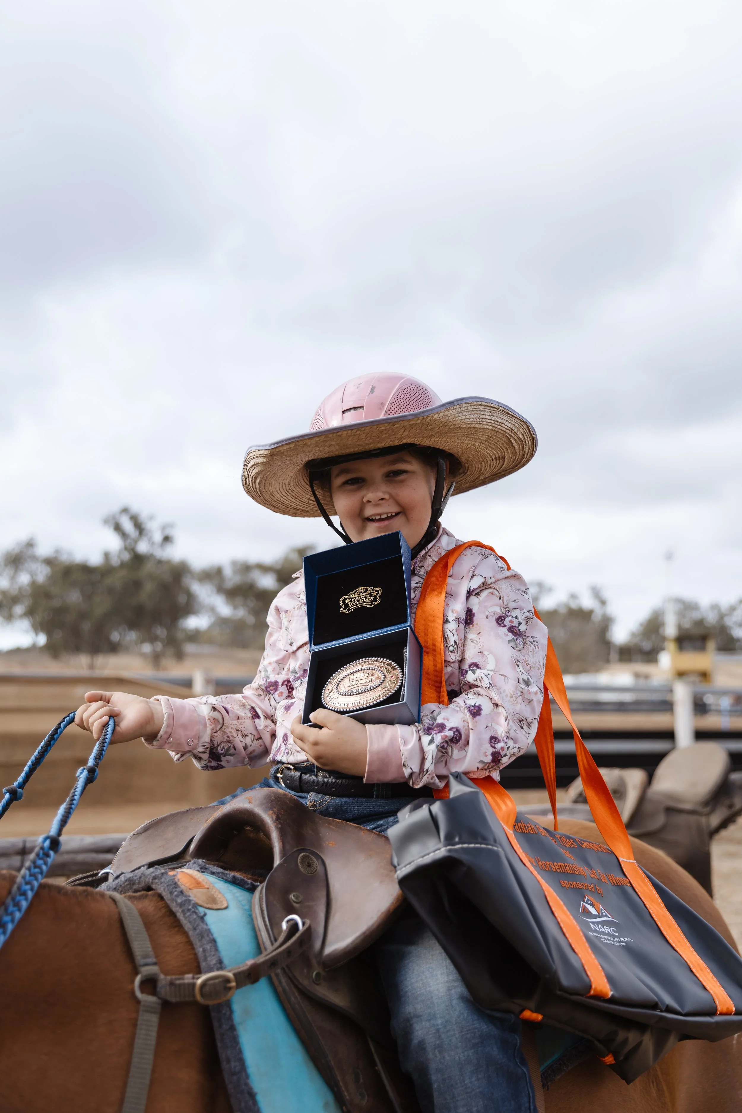A young girl sitting on a horse, wearing a large straw hat and holding an award medal in a box, outdoors on a cloudy day.
