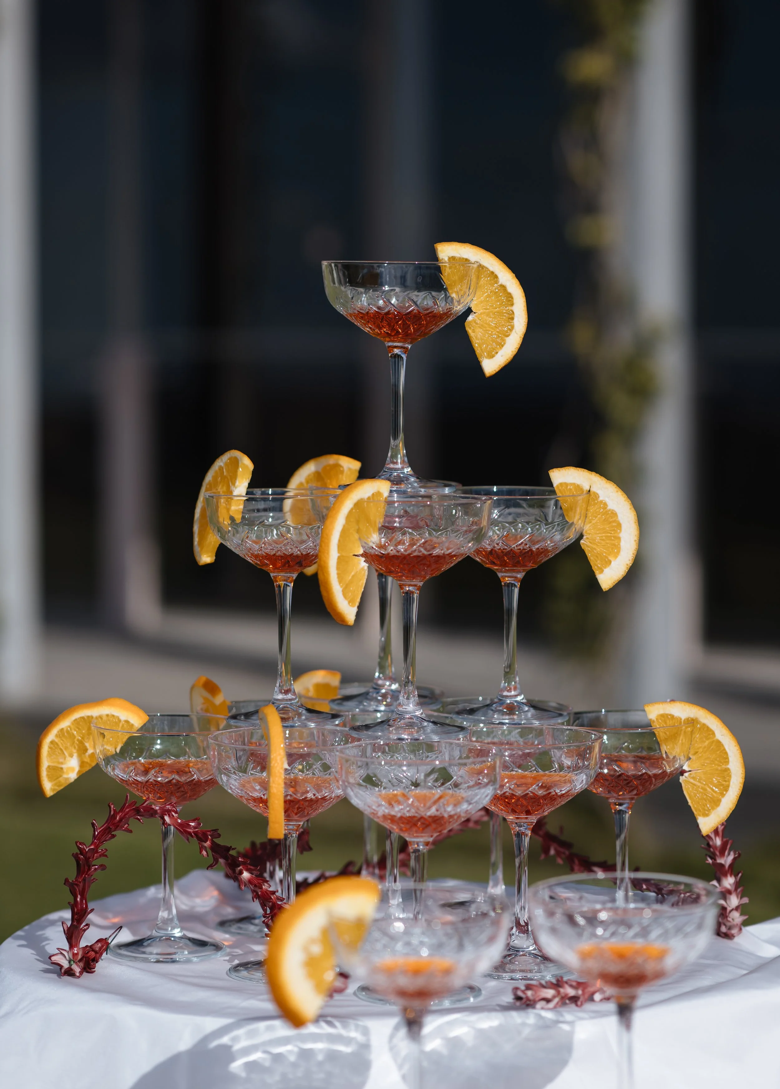 A multi-tiered glass stand holds several glasses filled with red drink, garnished with orange slices on the glasses' rims. The stand is outdoors with a dark background and some greenery, and the glasses are garnished with additional orange slices and