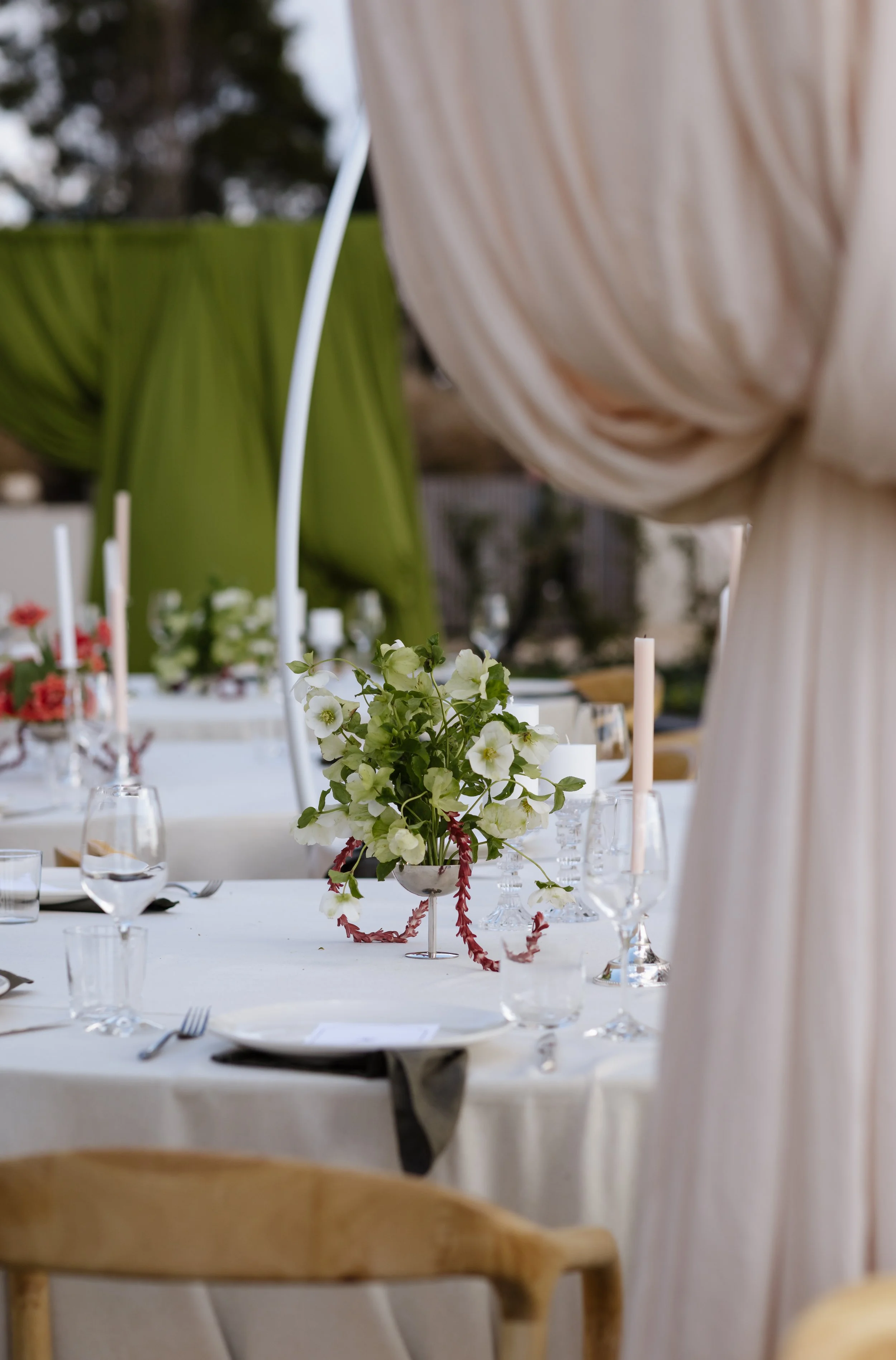 A decorated outdoor dining table with a white tablecloth, a floral centerpiece, candles, and glassware. Part of a beige curtain and a green backdrop are visible.
