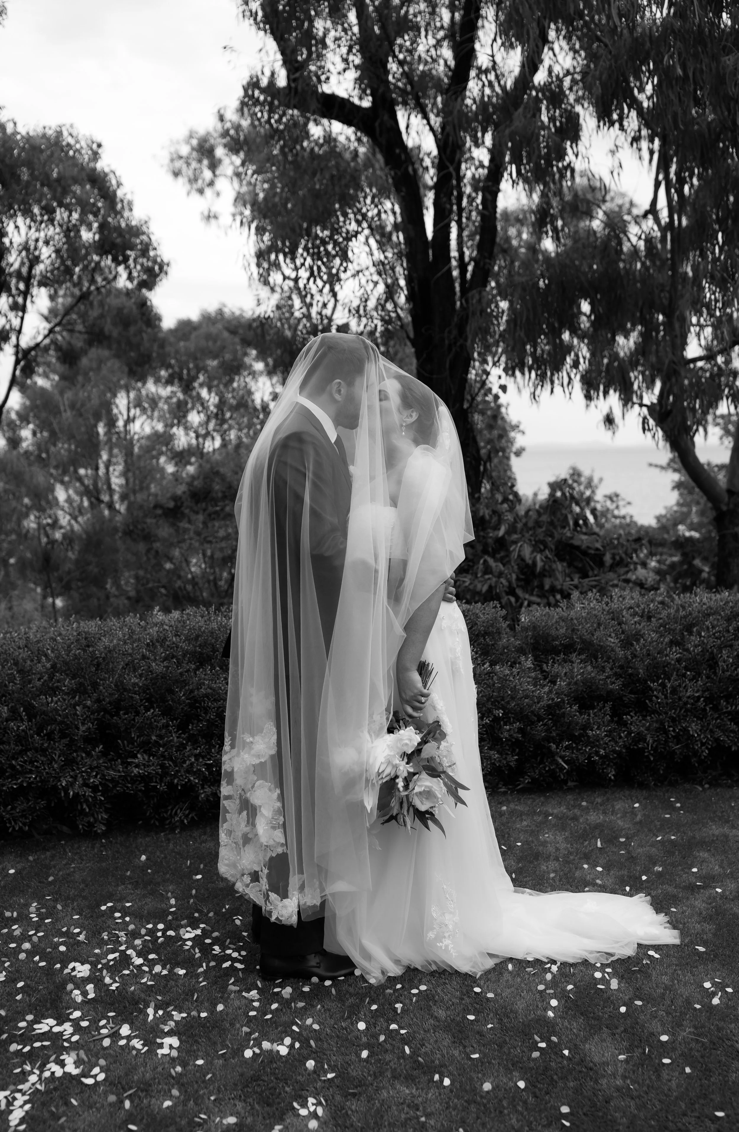 Black and white photo of a bride and groom standing close together outdoors, covered by a veil, sharing a kiss. The bride holds a bouquet, and flower petals are scattered on the grass around them, with trees and a body of water in the background.