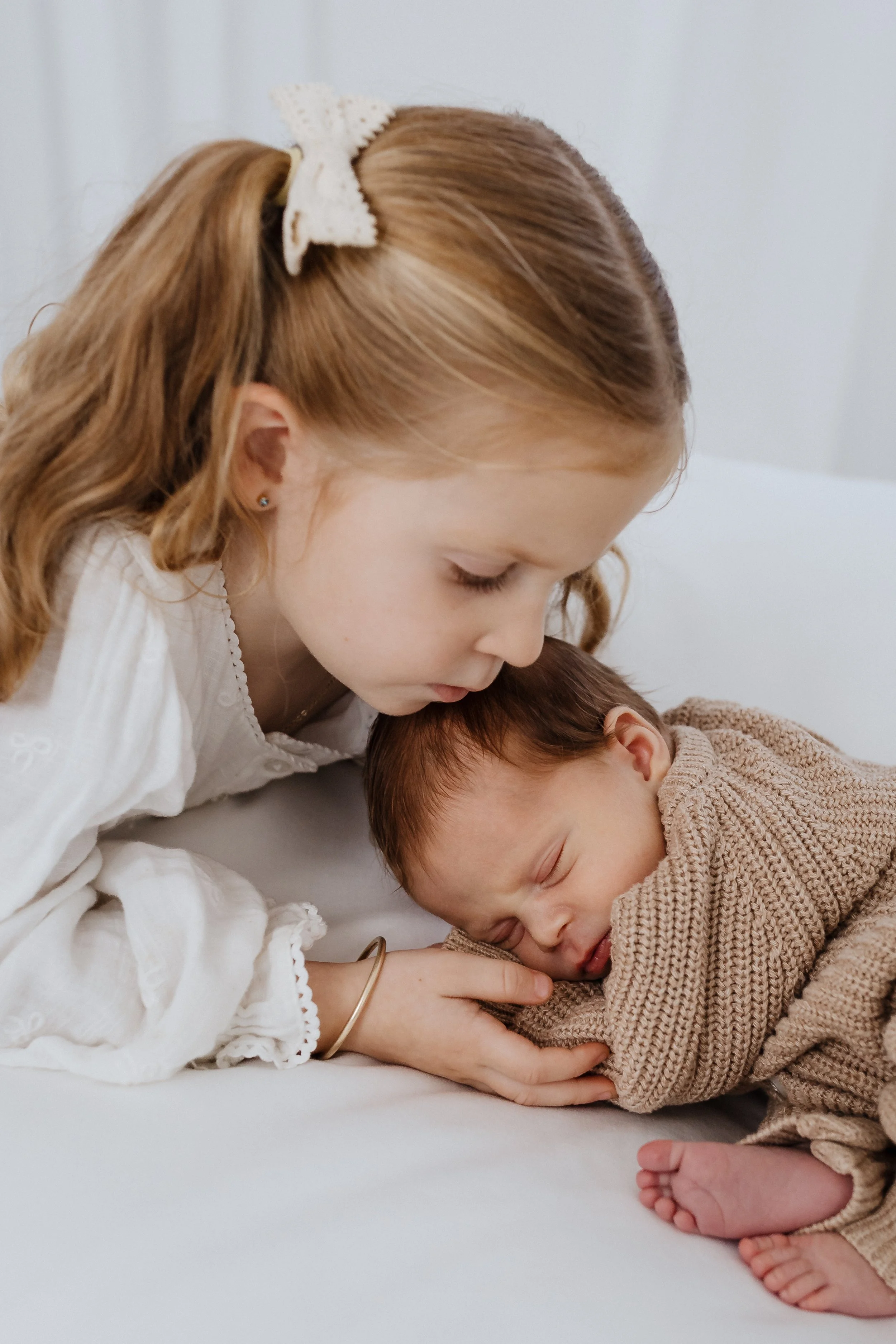 A young girl with red hair, a white bow, and earrings gently touches a sleeping baby with red hair and a beige sweater, both lying on a white surface.