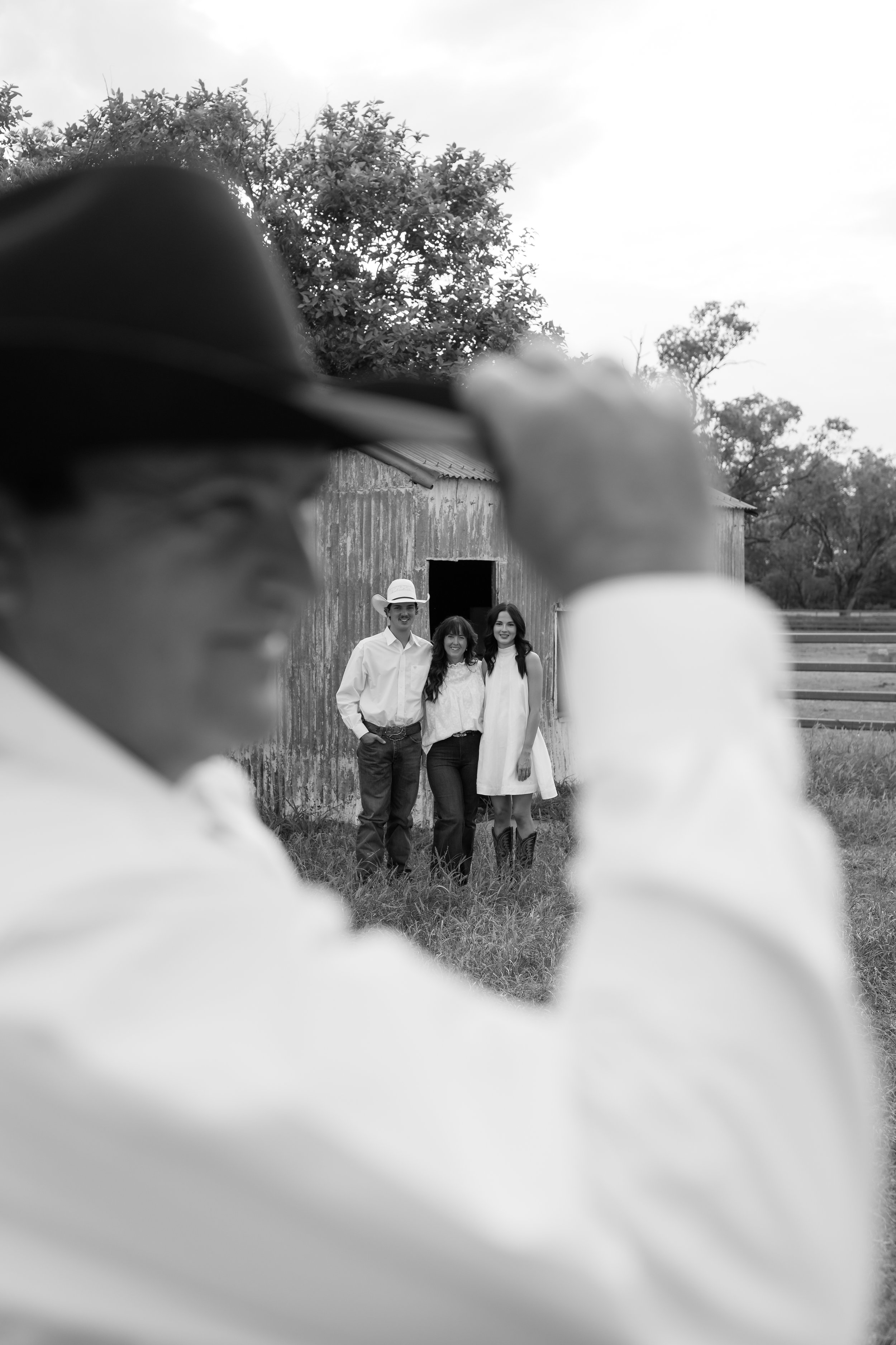 Black and white photo of three people standing outside near a barn, viewed through an out-of-focus person's shoulder and hat in the foreground. Two women and one man are smiling and standing close together, with trees and open land in the background.