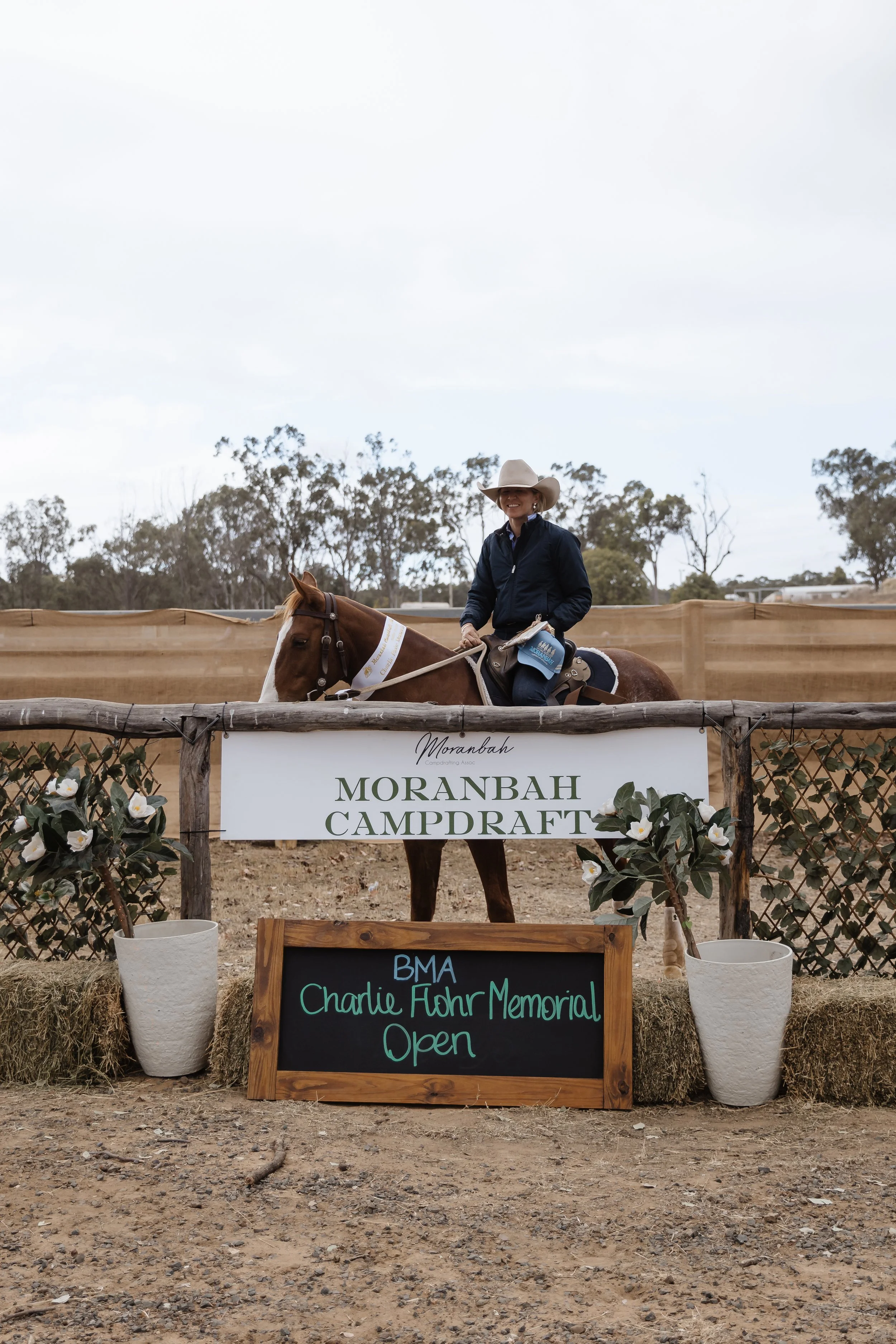 A woman wearing a cowboy hat, navy jacket, and jeans riding a brown and white horse on a dirt area. There are trees in the background and a sign that reads "MORANBAH CAMPROFT" in front of a decorative fencing with white flowers and vases. A smaller c
