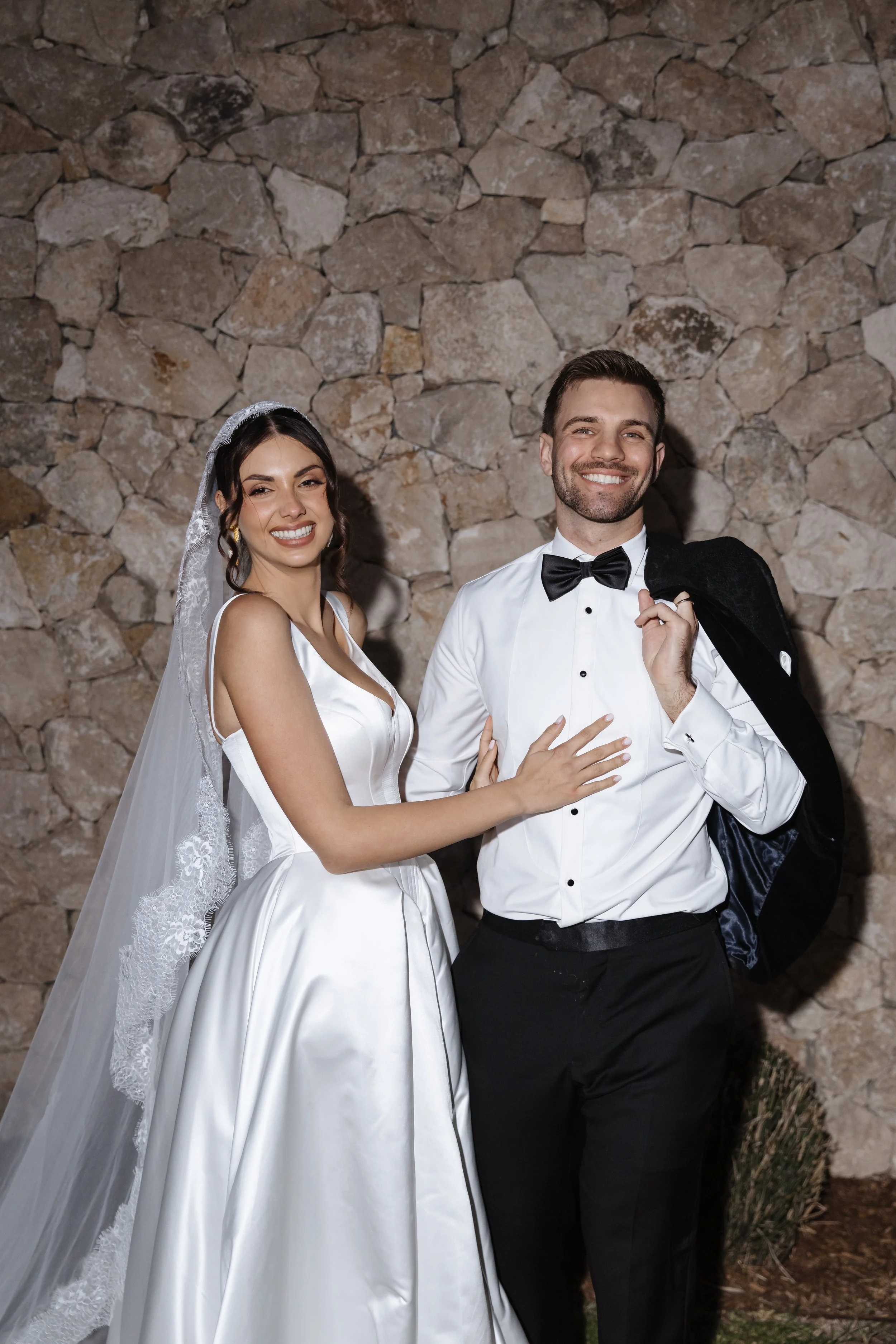 A bride and groom dressed in wedding attire, smiling against a stone wall background. The bride wears a white satin wedding gown with lace details and a veil, and the groom wears a white dress shirt with a black bow tie and is holding a black jacket 