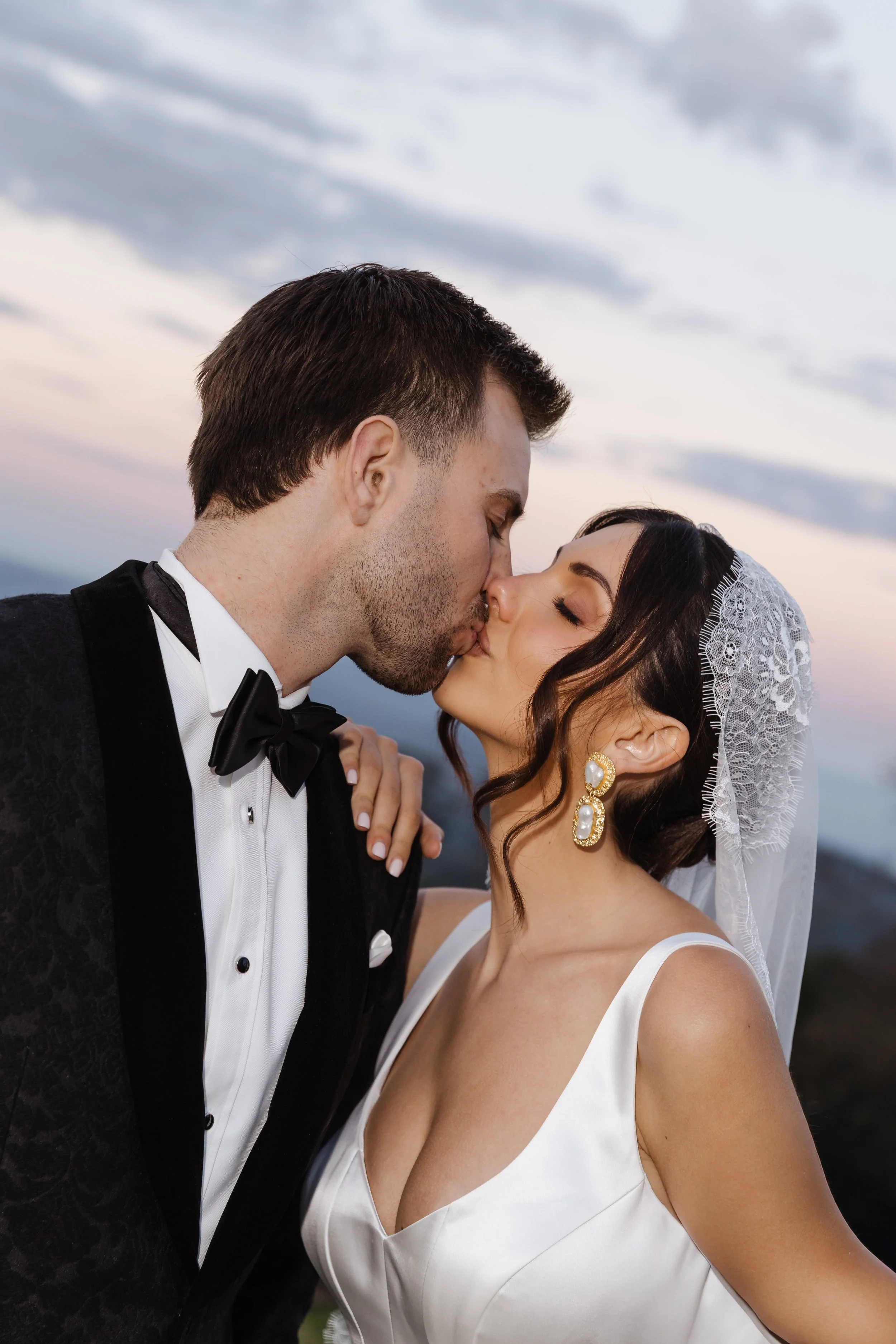 A bride and groom share a kiss outdoors during sunset, with the bride wearing a white dress and lace veil, and the groom in a black tuxedo.