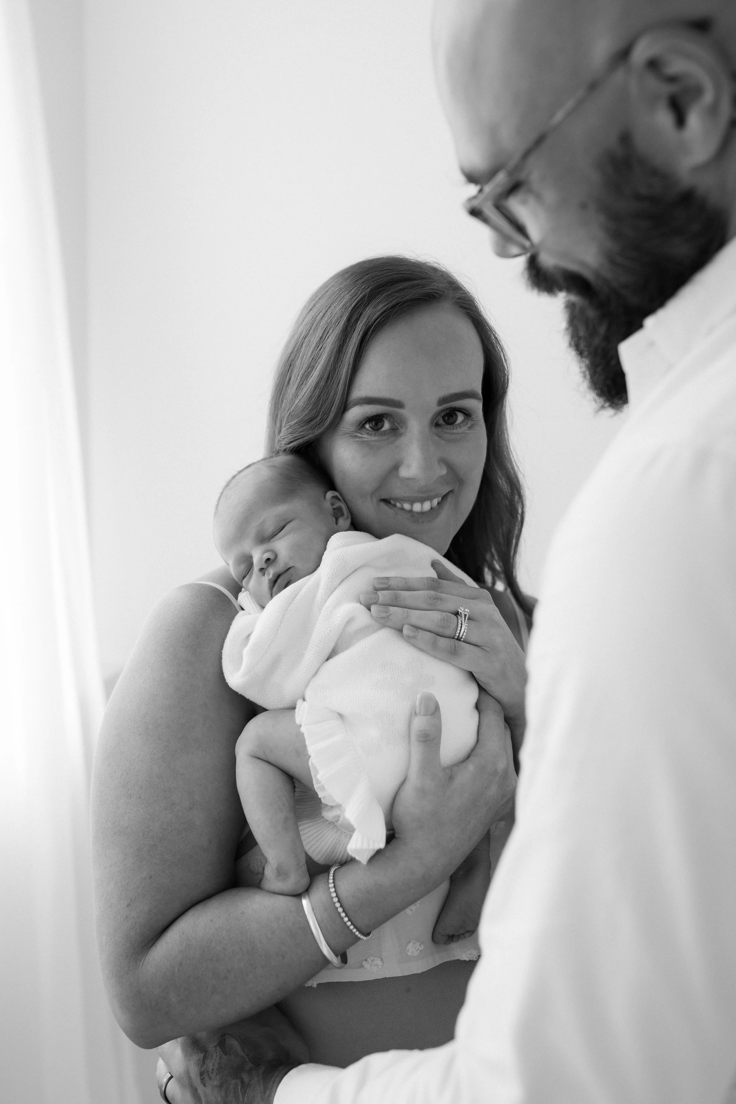 A woman holding a newborn baby, smiling, while a man wearing glasses and a white shirt looks at them, in a hospital setting.