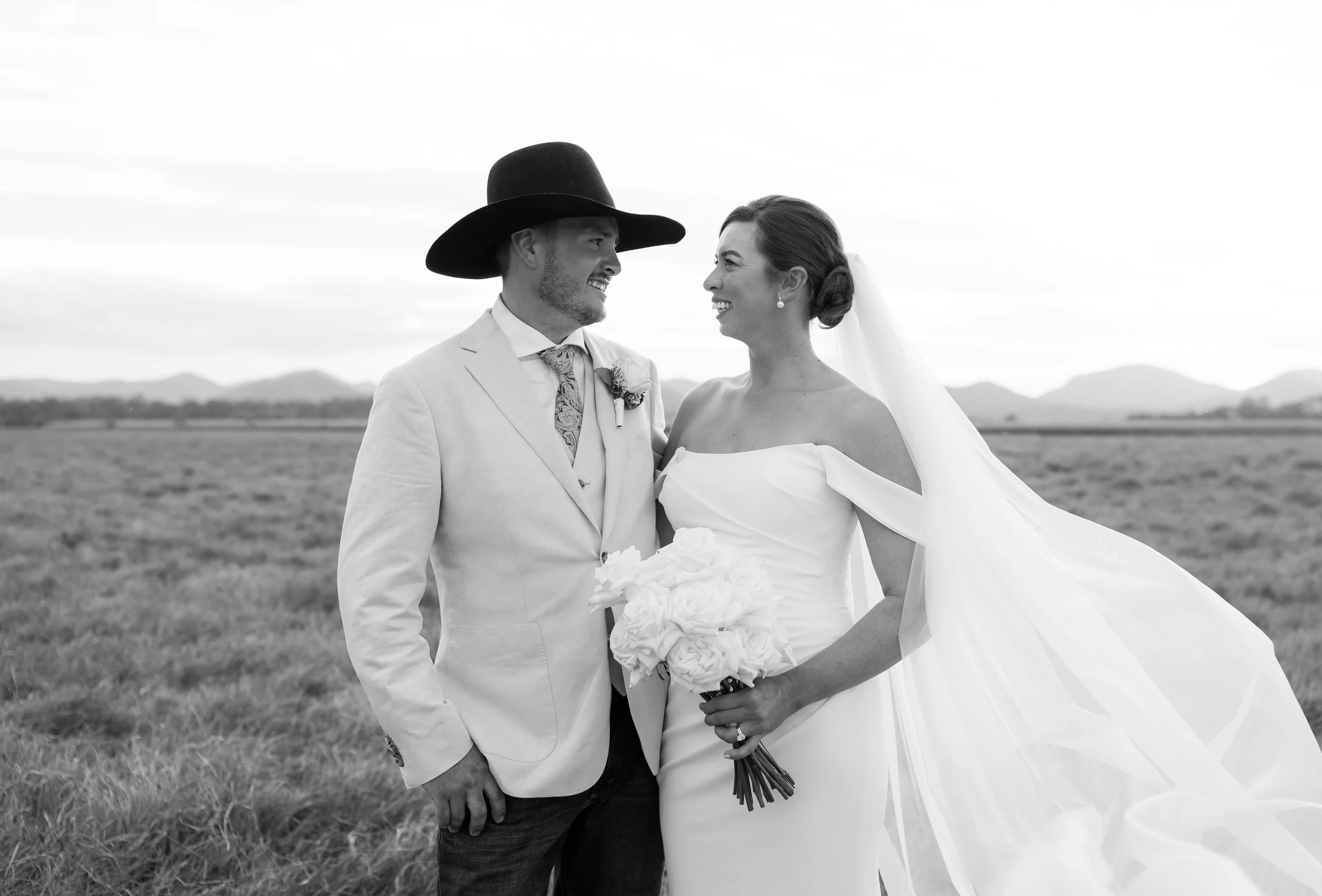 Black and white photo of a bride and groom standing outdoors in a field. The groom, wearing a light-colored suit and large hat, looks at the bride. The bride, in an off-the-shoulder wedding dress holding a bouquet of flowers, smiles back at the groom