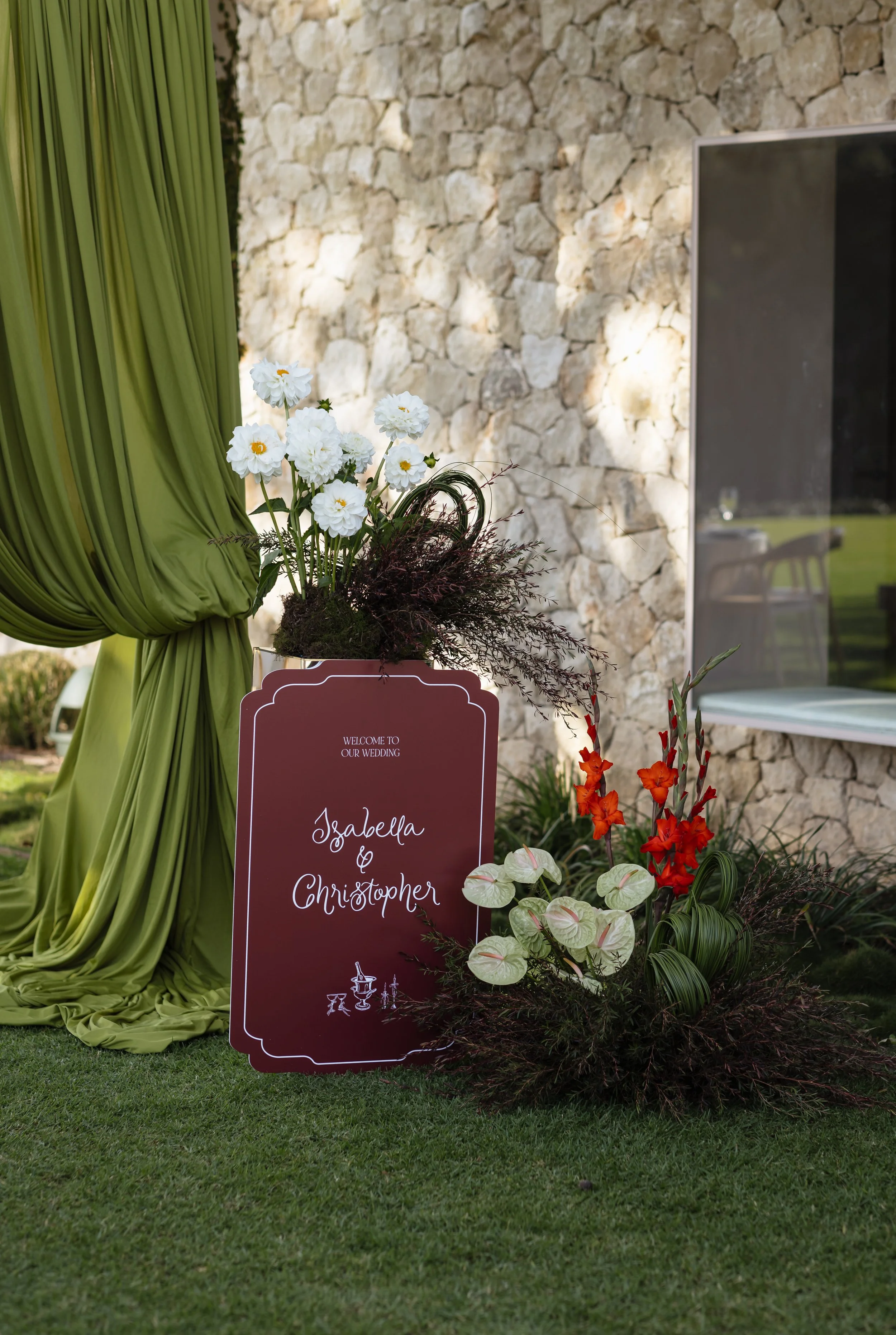 A wedding sign with the names Isabelle and Christopher, decorated with white flowers, orange flowers, and green foliage, placed outdoors in front of a stone wall, next to a green curtain.