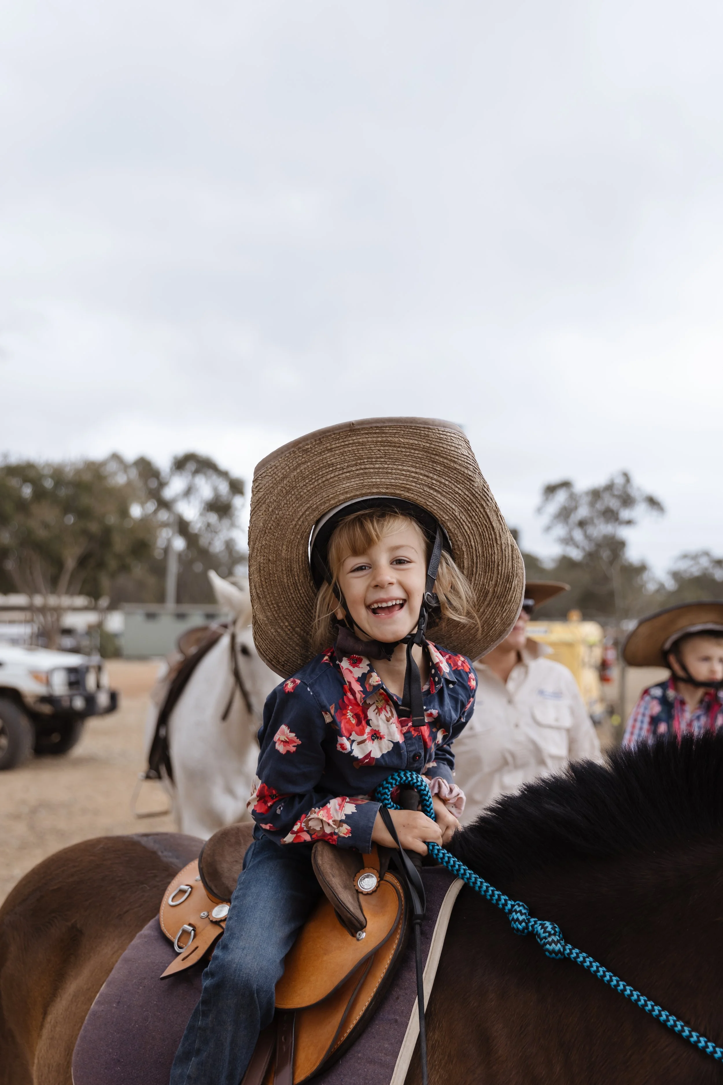 A young girl with blonde hair and a big smile riding a horse, wearing a large straw hat and a navy blue floral shirt, with other children and trees in the background.