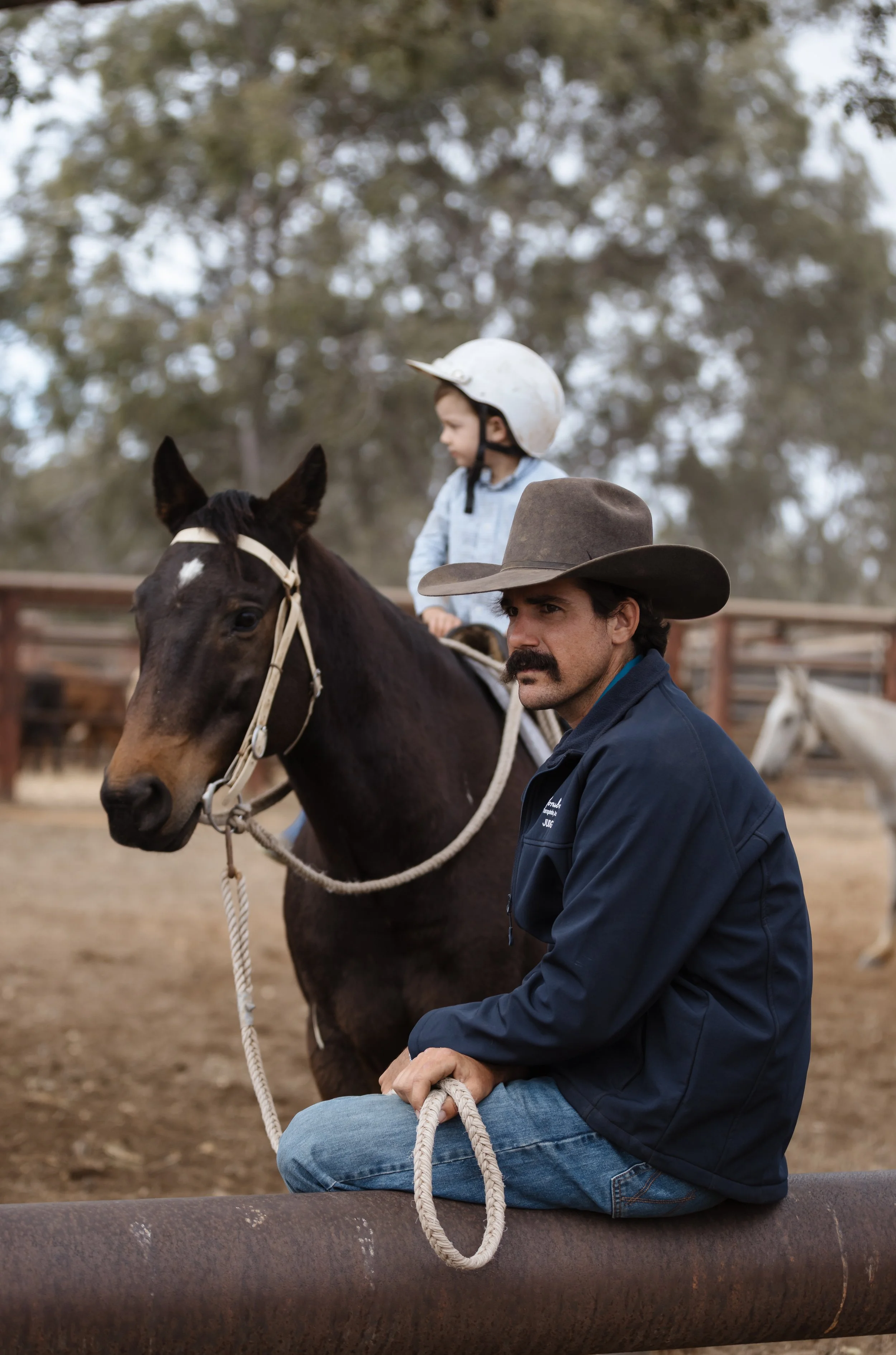 A man with a mustache and beard sitting on a horse, wearing a wide-brimmed hat and a blue jacket. A young boy wearing a bike helmet is sitting on the same horse, looking ahead. They are at a riding facility with a wooden fence and trees in the backgr