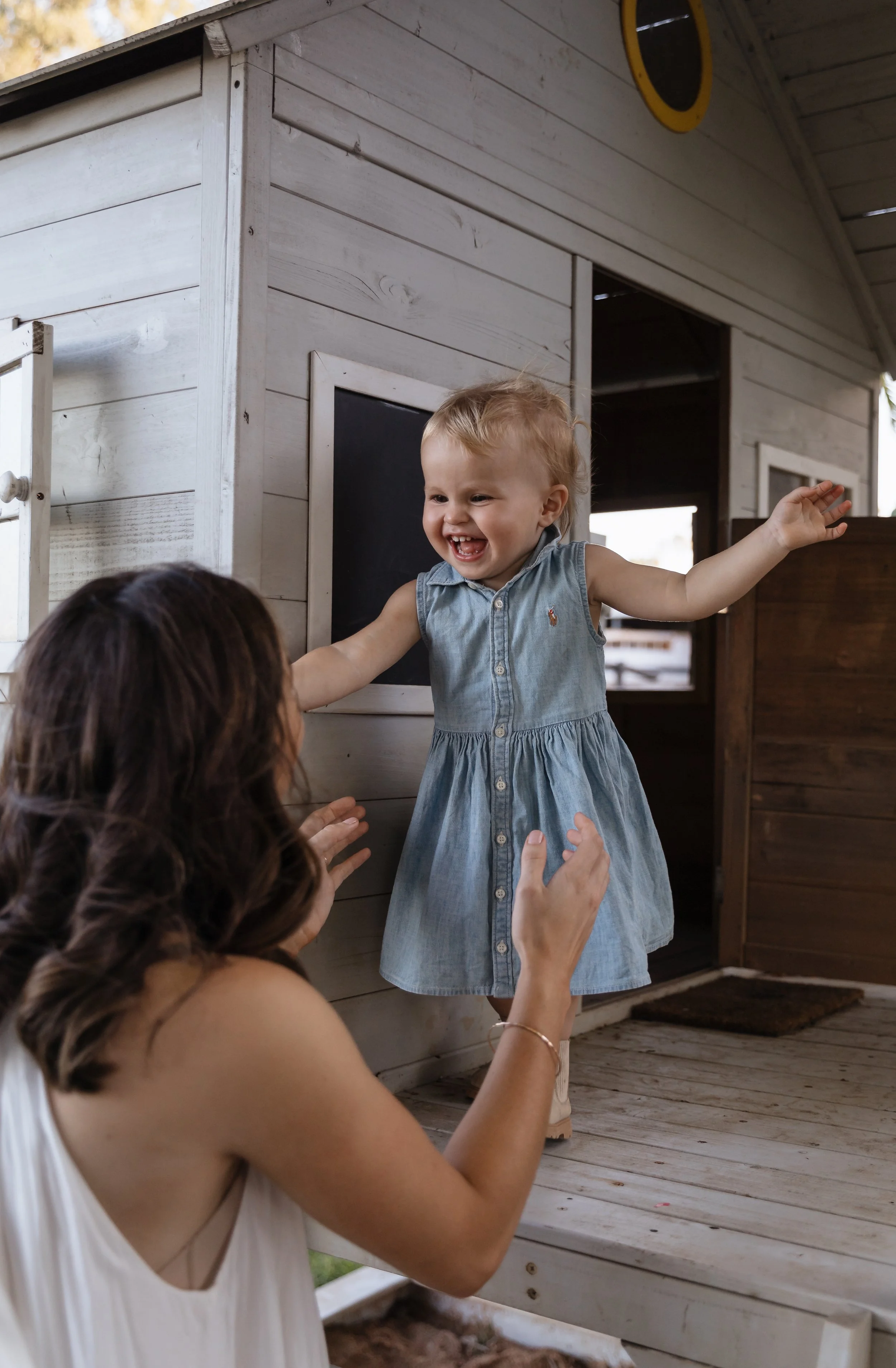 A young girl in a sleeveless denim dress with embroidered emblem smiling and reaching out her arms toward a woman with long, wavy brown hair, who is raising her hands. They are outside near a small wooden playhouse with a black chalkboard and a windo