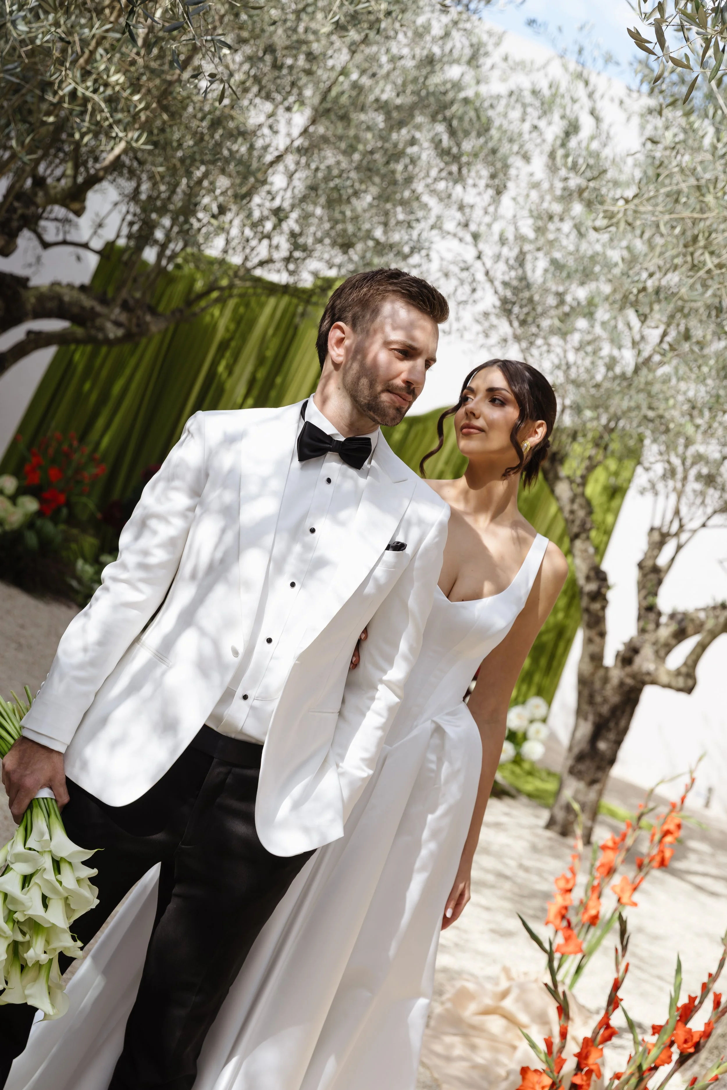 A couple dressed in wedding attire standing outdoors under trees with dollops of sunlight and shadows, with flowers and greenery around them.