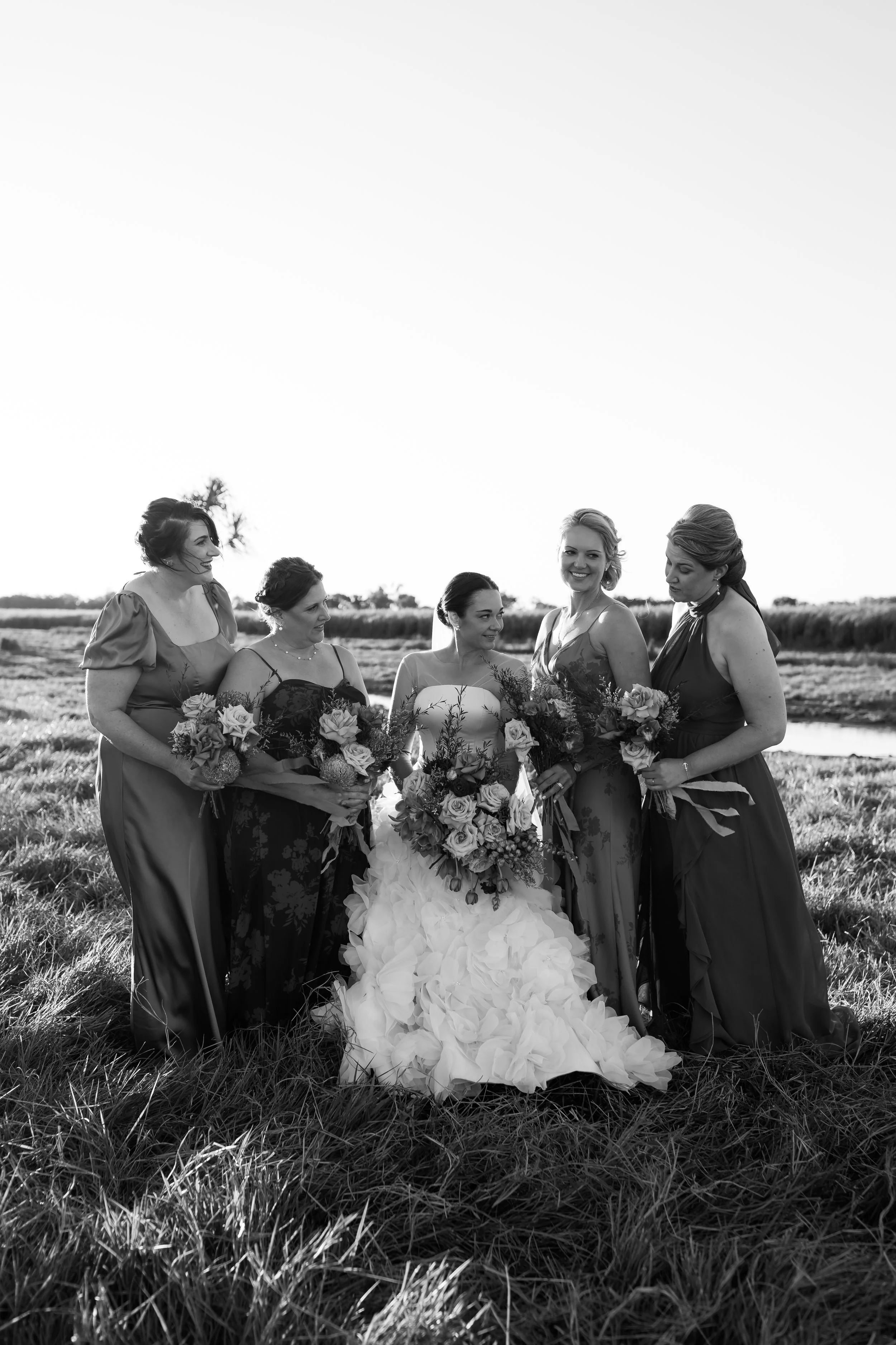 Black and white photograph of six women in formal dresses standing outdoors in a field, holding bouquets of flowers, with a clear sky and horizon in the background.