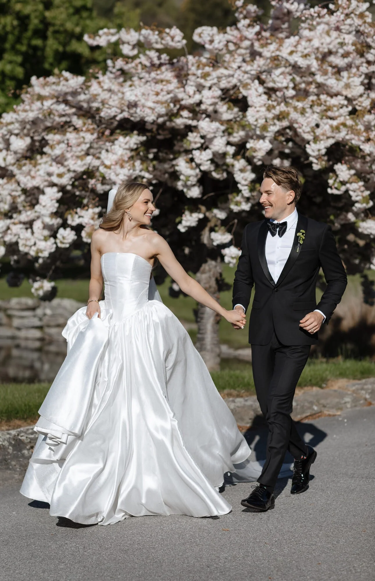 A bride and groom holding hands and smiling, walking outdoors in front of a flowering tree with pink and white blossoms.