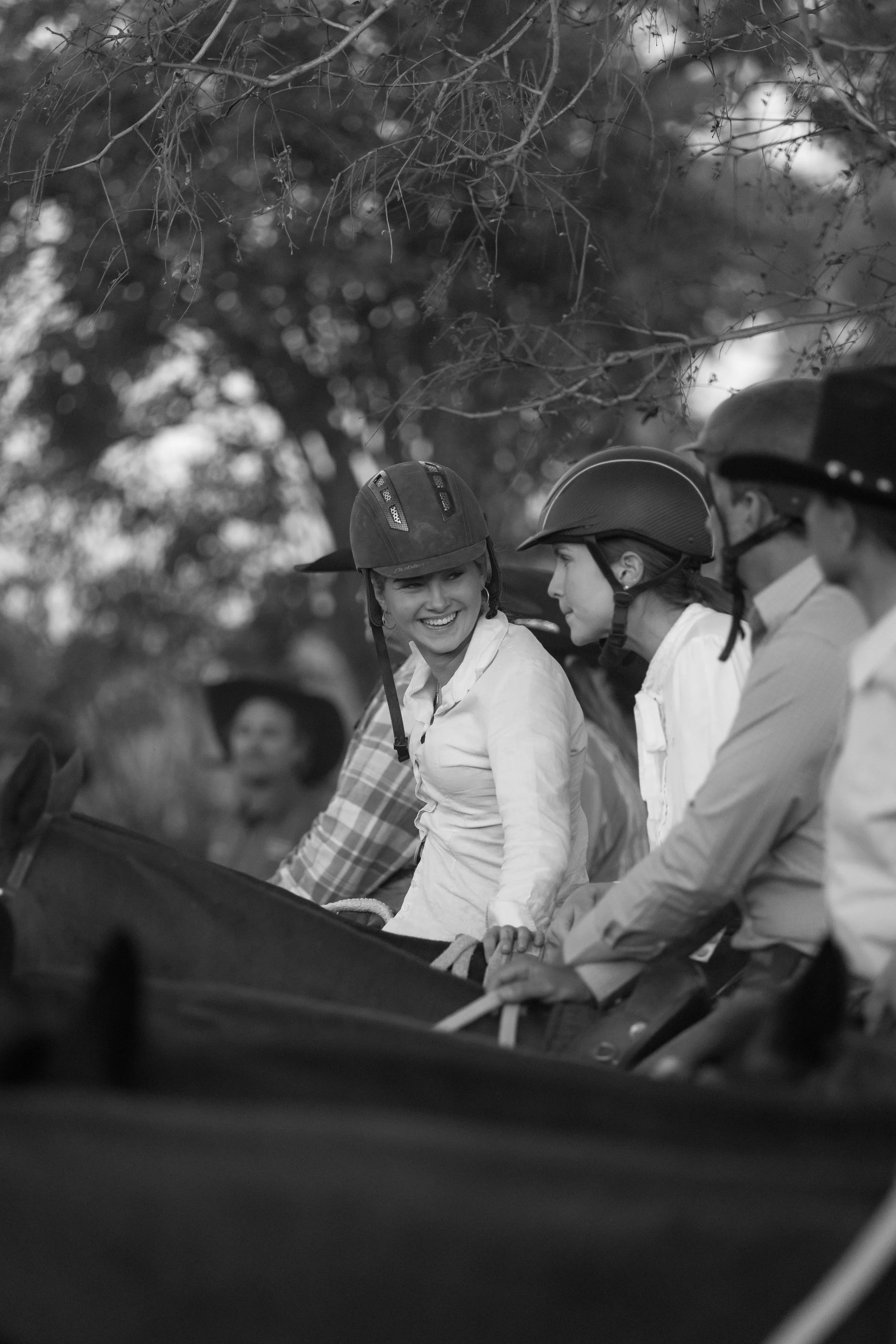 Four people, wearing helmets, are sitting on a horse-drawn carriage outdoors, engaging in a conversation and smiling.