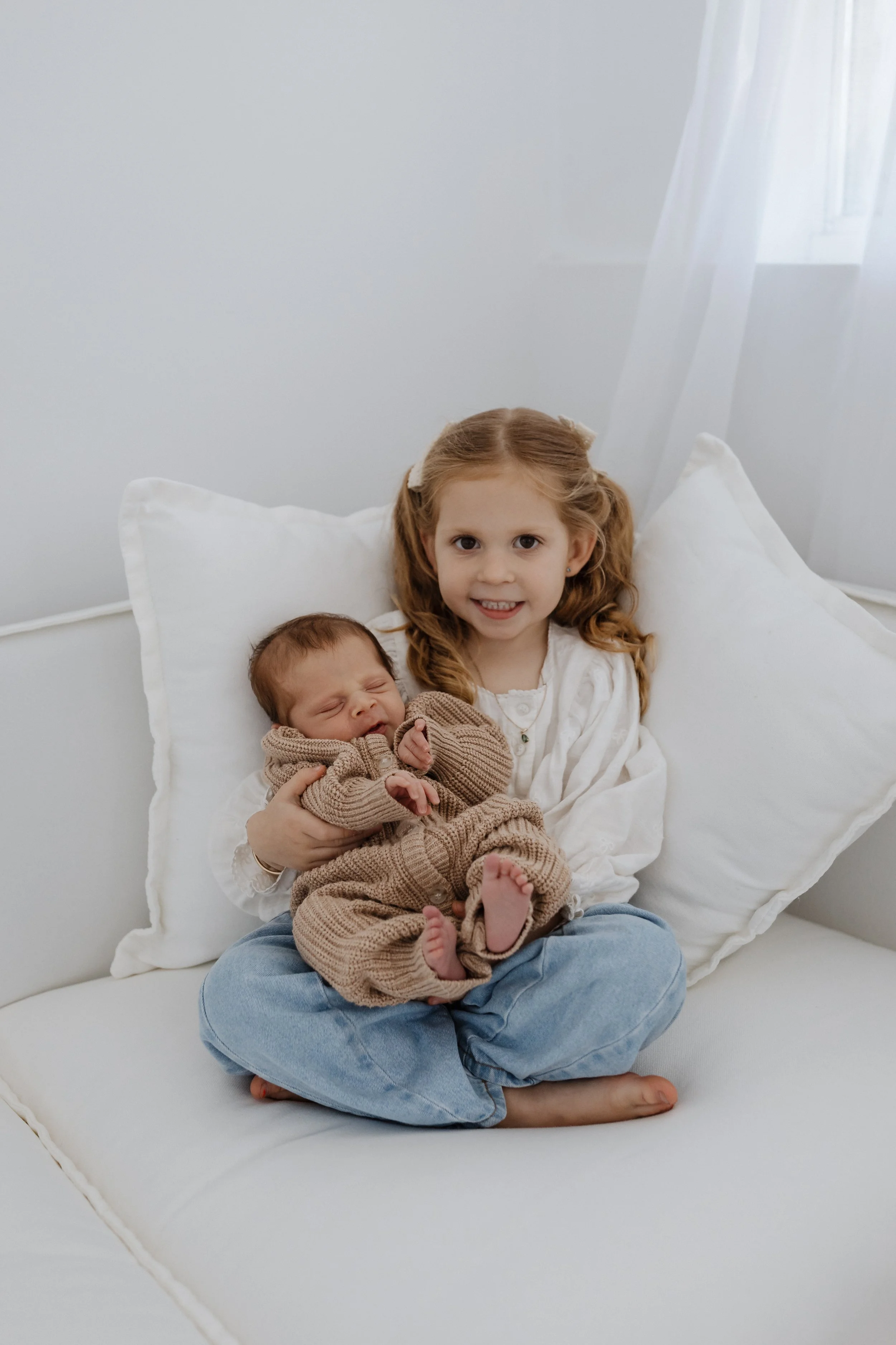 A young girl with curly red hair holding a sleeping baby in her lap on a white couch