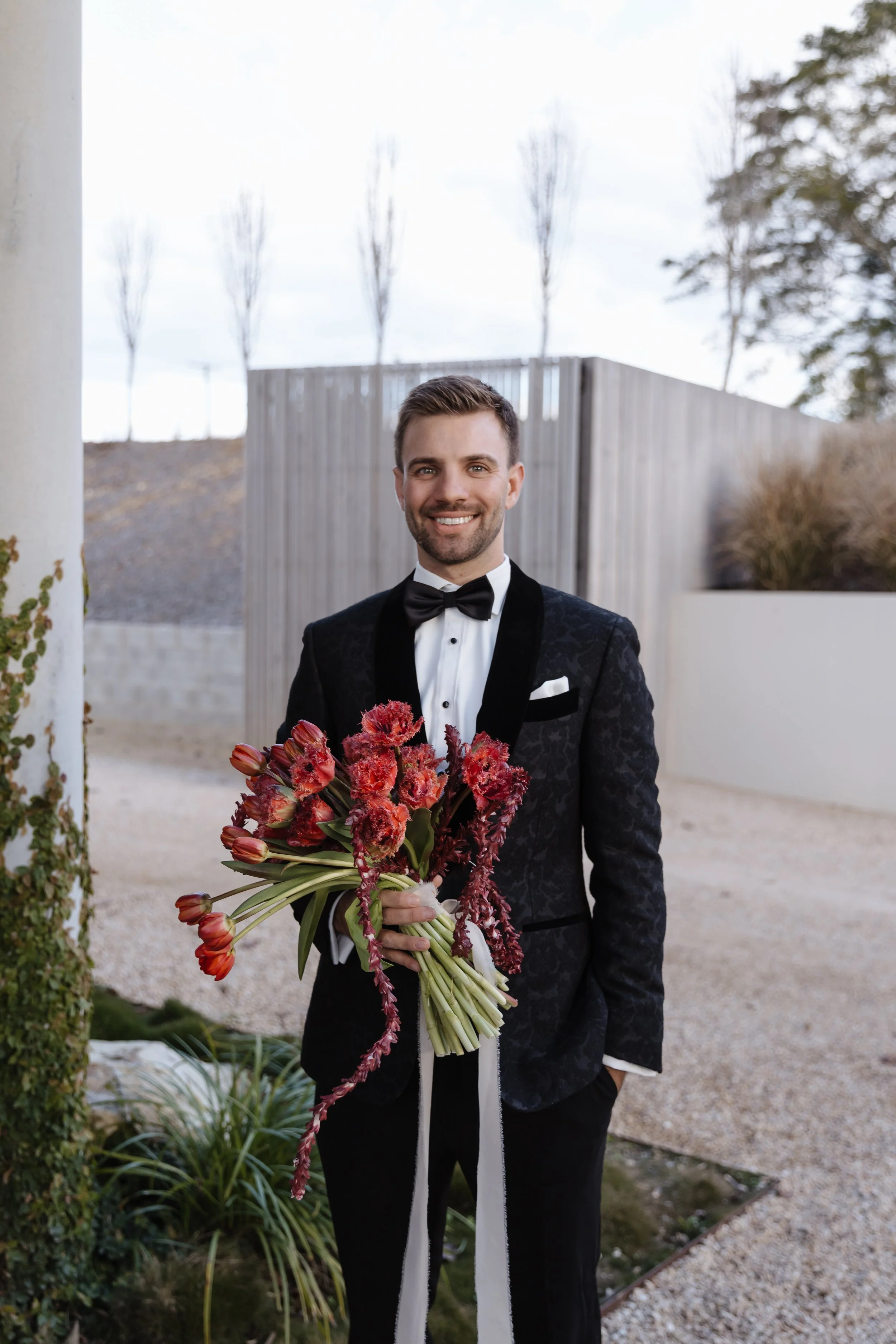 A man dressed in a black tuxedo with a bow tie standing outdoors, holding a large bouquet of red and pink flowers with long green stems, smiling at the camera.