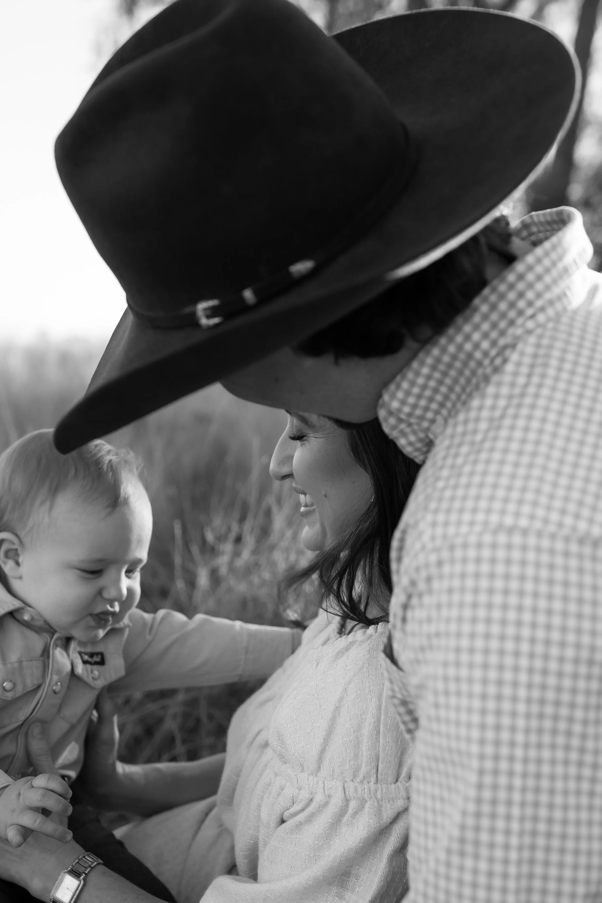 A woman with long dark hair, wearing a light-colored textured sweater, is smiling and facing a young boy. The woman is wearing a wide-brimmed hat and a checkered shirt. The boy, with short hair, is reaching and touching the woman's arm. The photo is 
