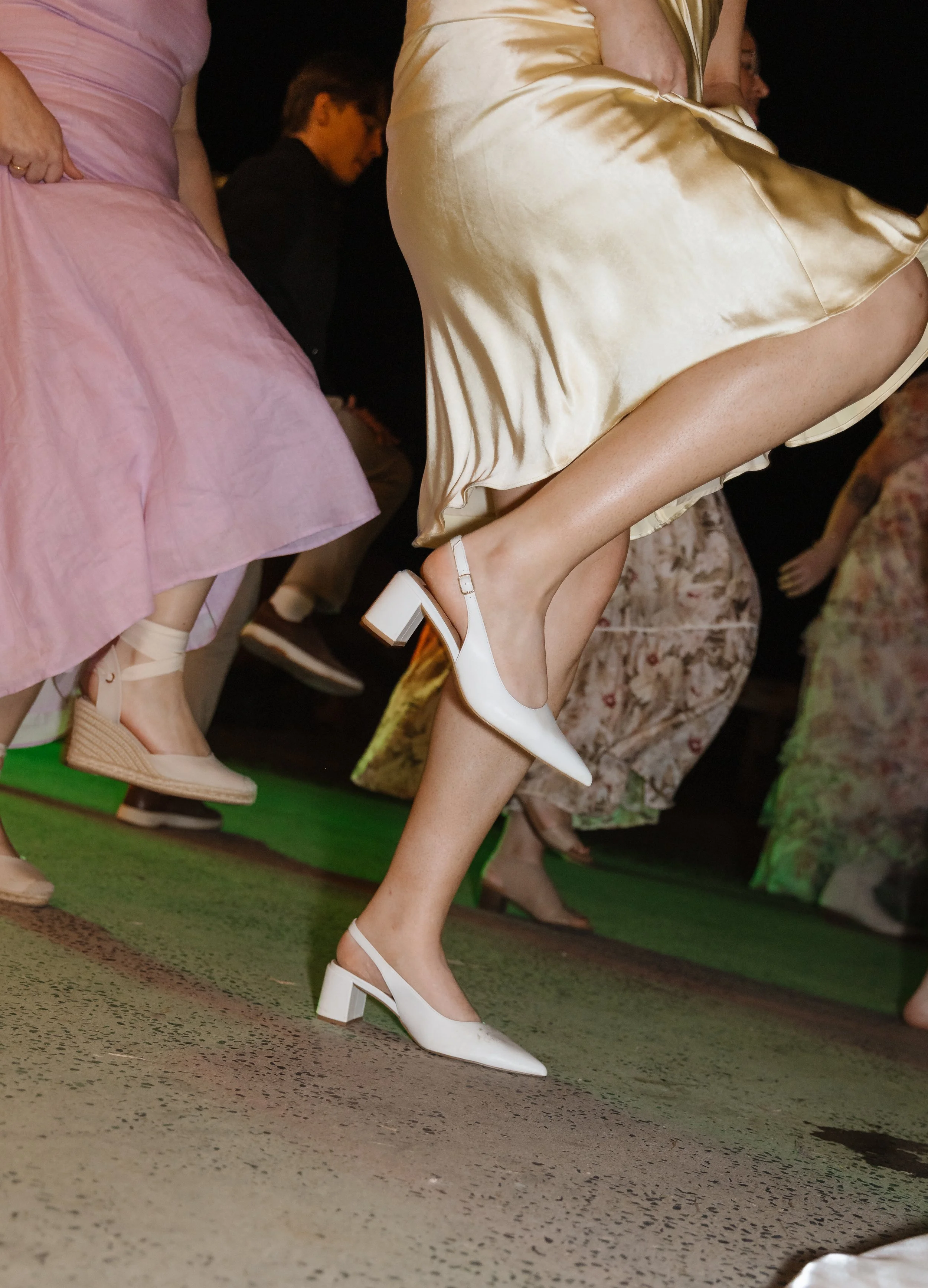 Close-up of women in vintage dresses and high heels, with focus on their legs and footwear, standing on a patterned carpet at an indoor event.