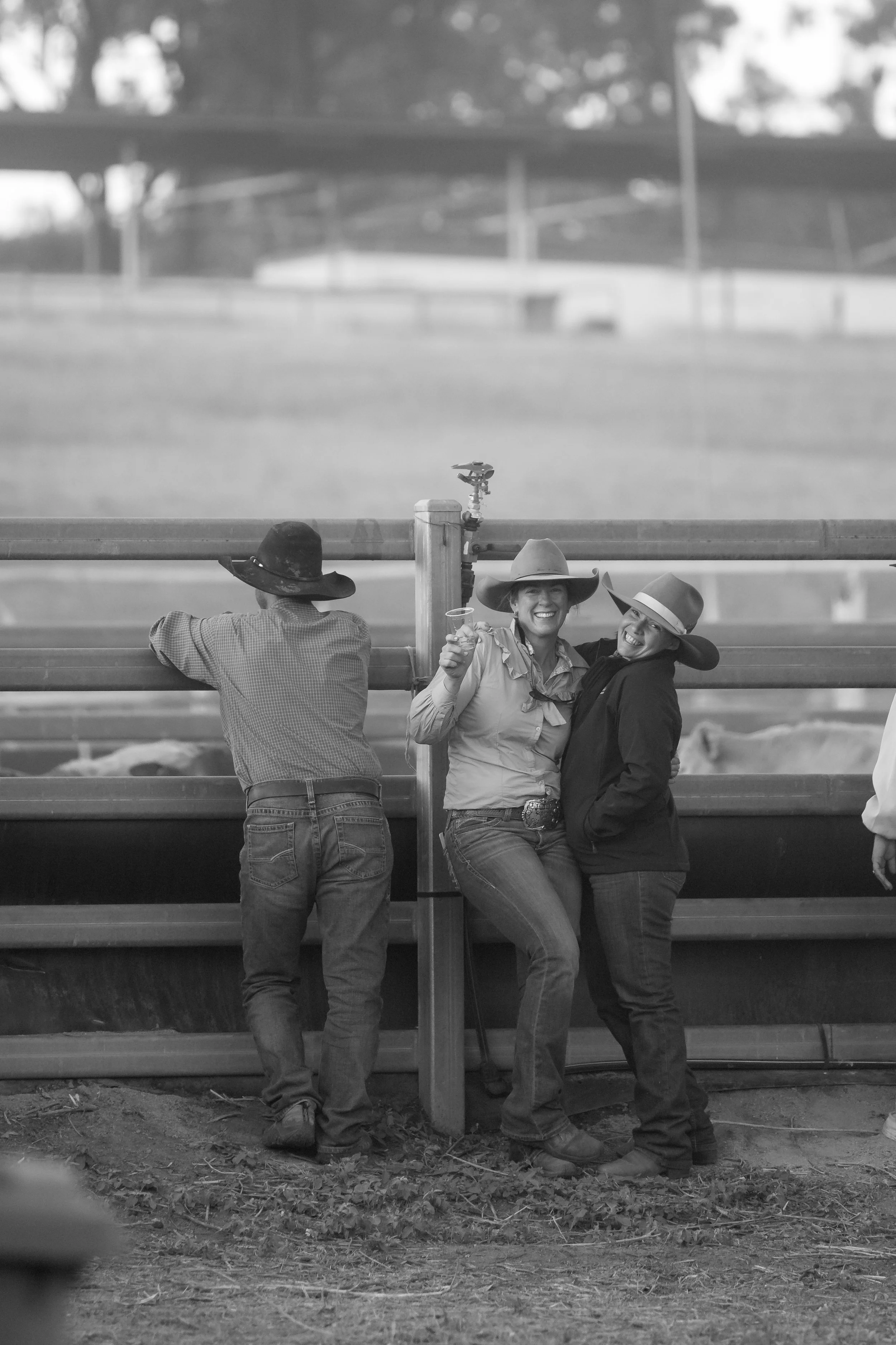 Three people in cowboy hats are seen sitting and standing behind a fence at a rodeo event, smiling and posing for the photo.
