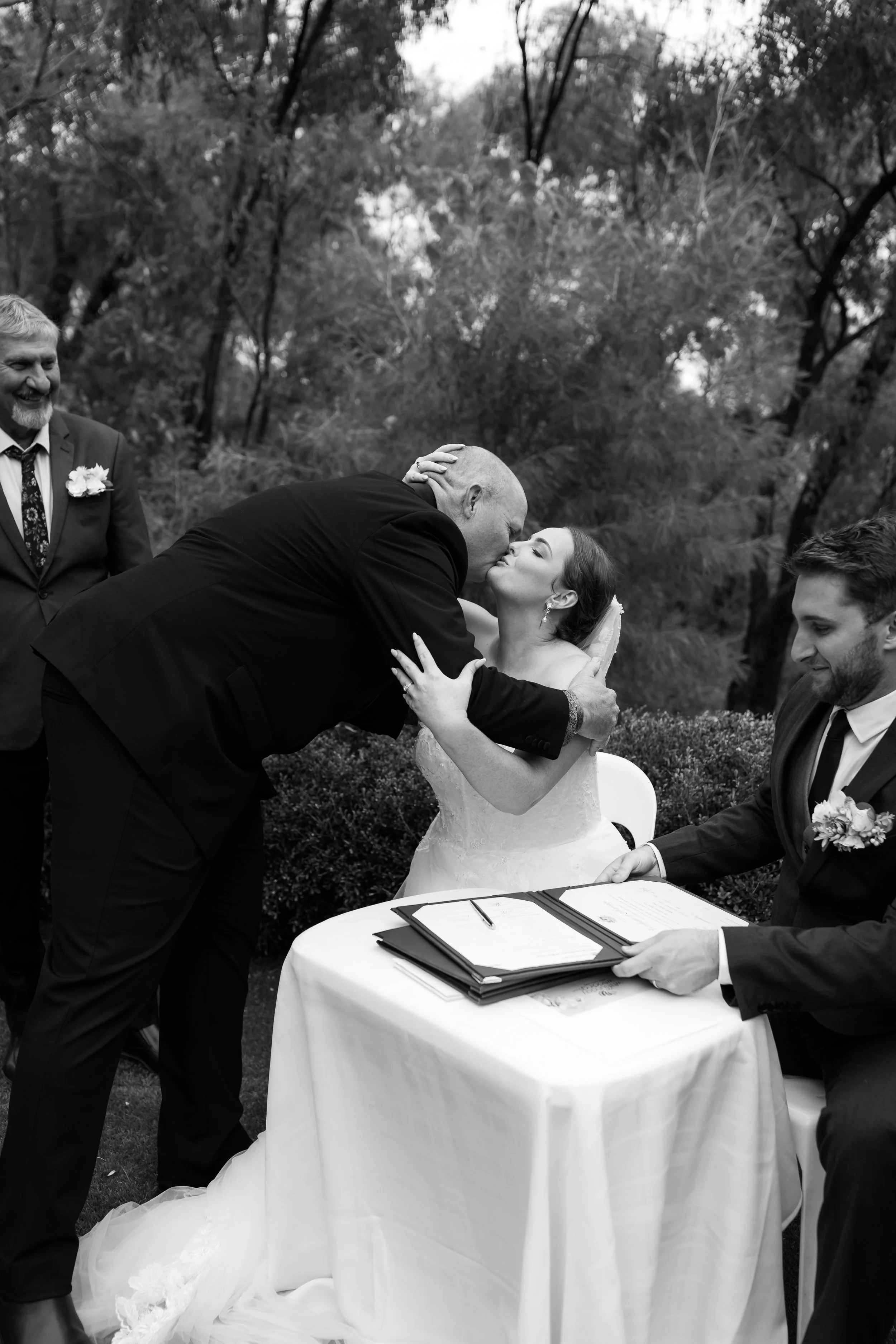A black and white photo of a wedding ceremony outdoors. The bride and groom are kissing, with the groom bent over the bride, who is seated. An officiant is holding an open book or folder on a table in front of them. Two other men are visible, one smi