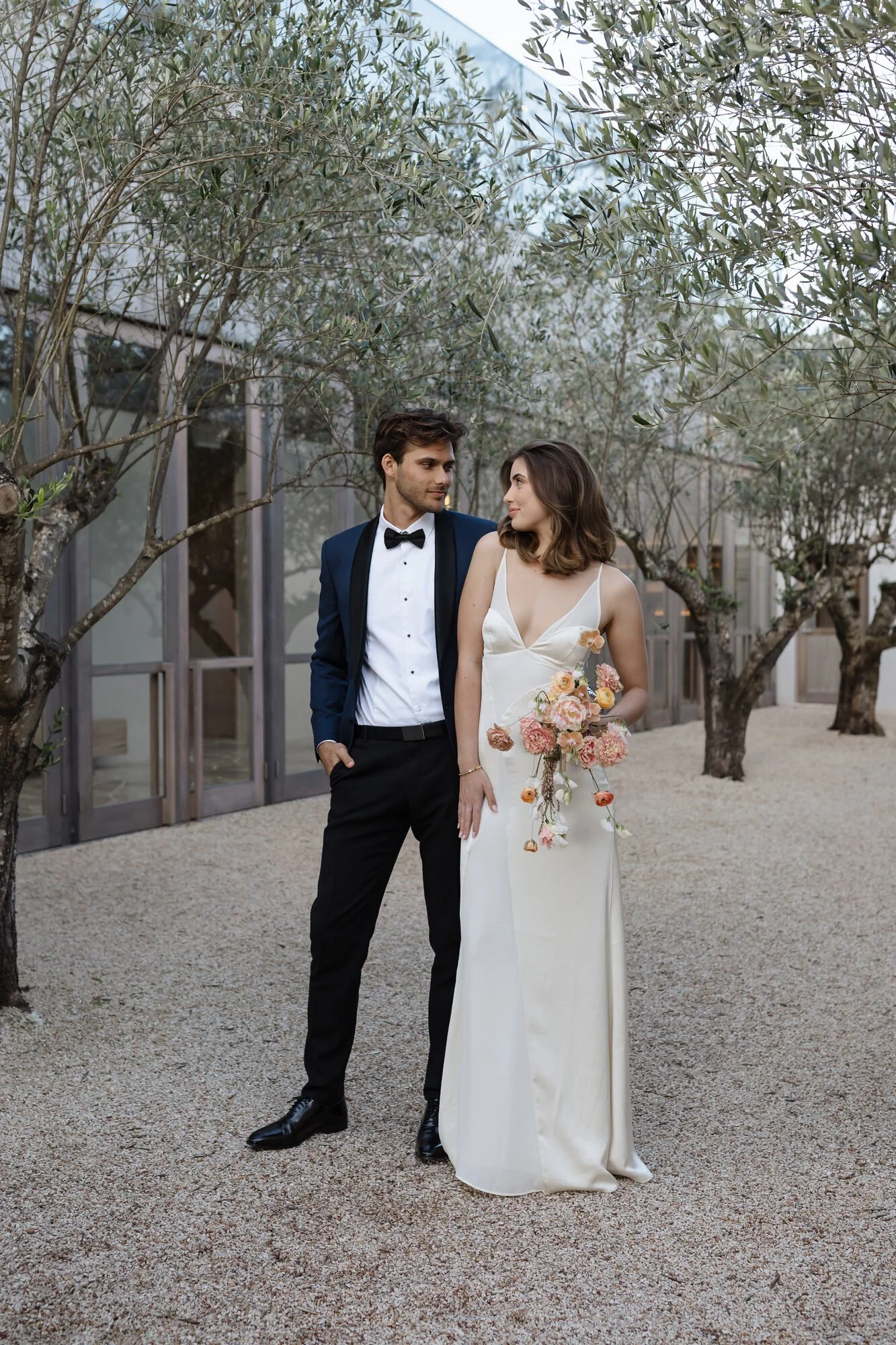 A young man and woman dressed in formal wedding attire standing outdoors under trees with a modern building in the background. The man is wearing a navy tuxedo with a bow tie, and the woman is in a white dress holding a bouquet of pink and orange flo