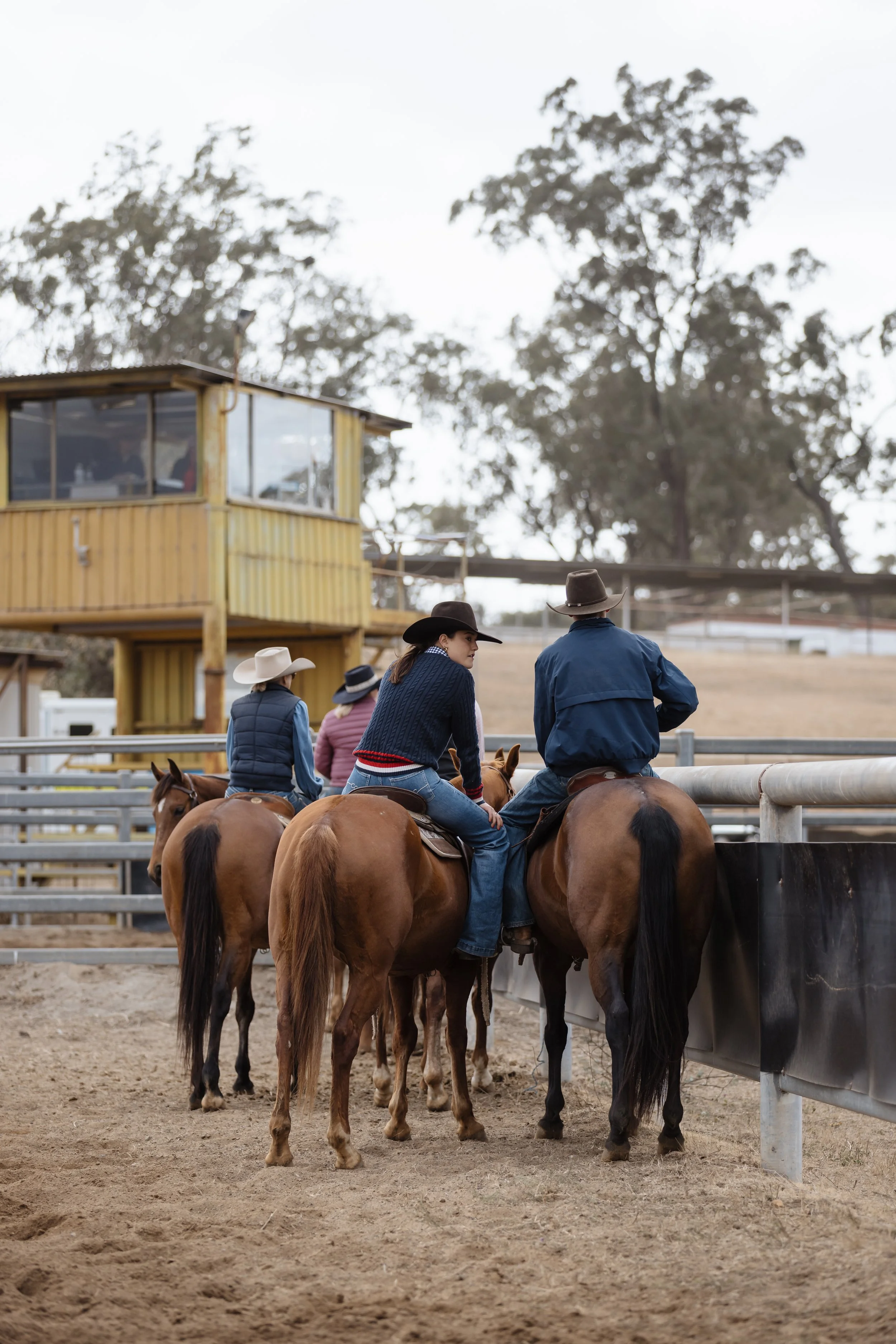 People riding horses at a rodeo or western event, with some wearing cowboy hats, on a dirt arena with a yellow wooden building and trees in the background.