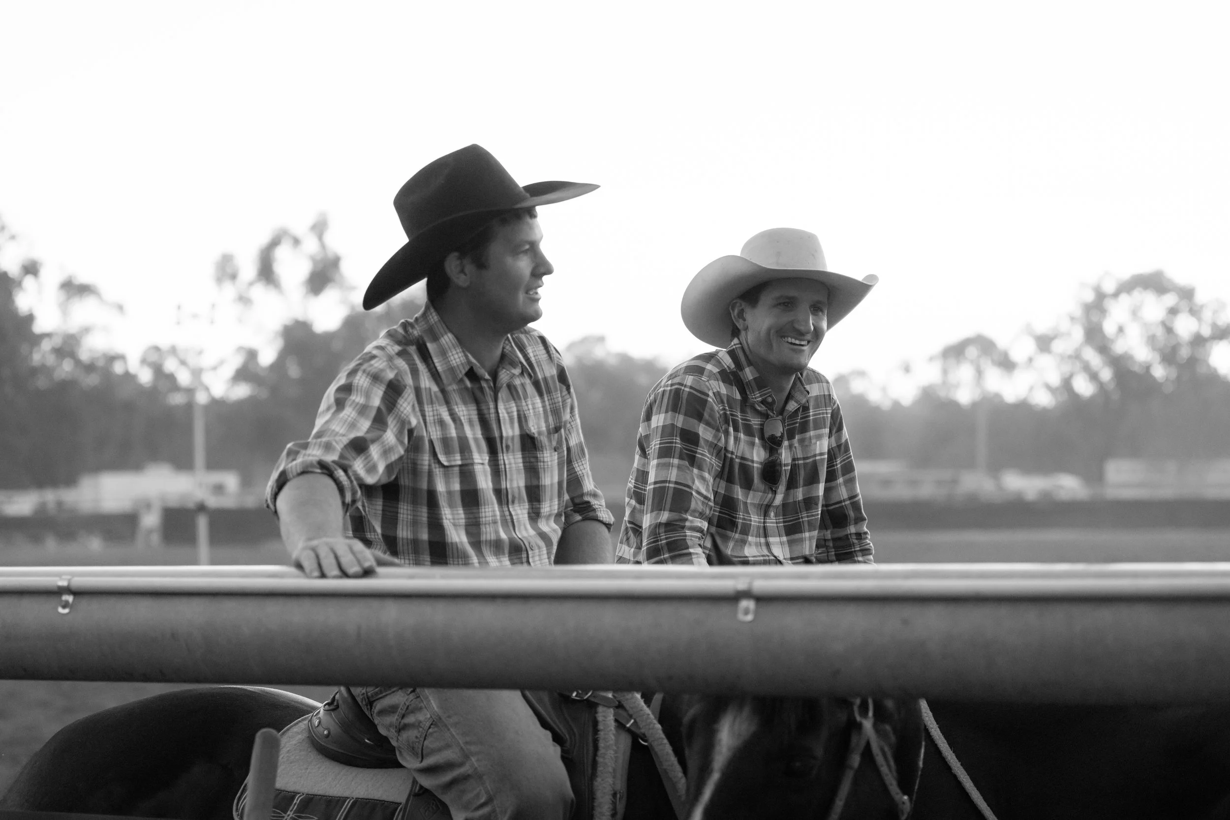 Two men in cowboy hats riding a horse in a rural outdoor setting, wearing plaid shirts, with trees and open sky in the background.