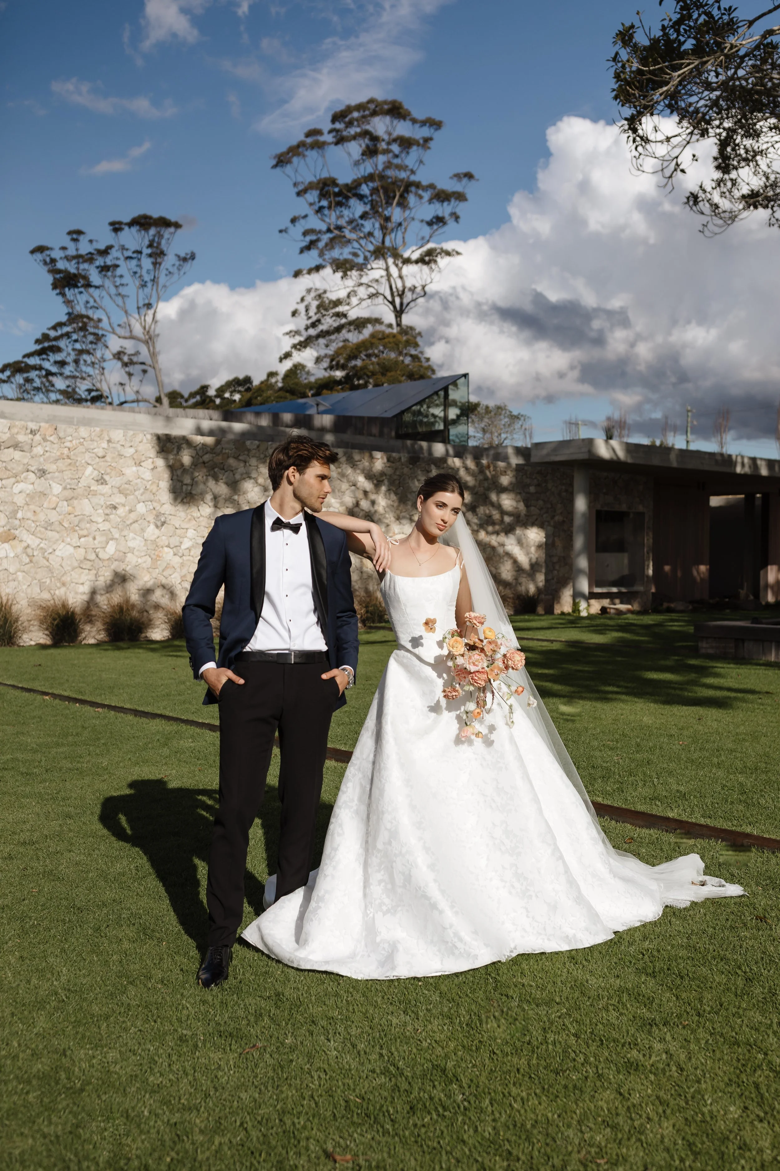 A bride and groom standing outdoors on a grassy area during daytime. The groom is wearing a dark suit with a bow tie and has his hands in his pockets. The bride is wearing a white wedding gown with a long train, holding a bouquet of flowers, and look