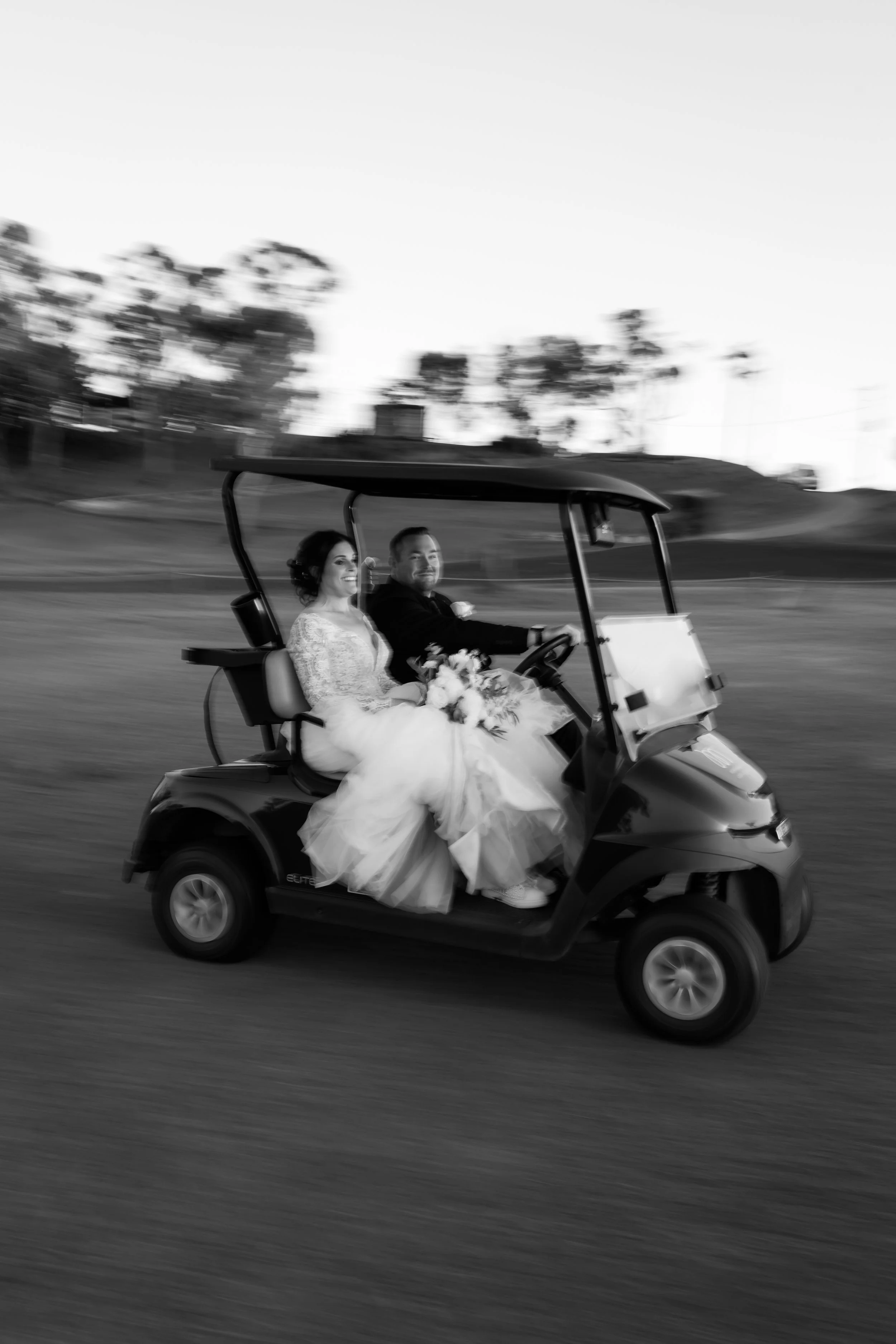 A black and white photo of a newlywed couple riding in a golf cart. The bride, wearing a wedding dress, holds a bouquet and is smiling, while the groom, in a suit, is driving and also smiling. The background is blurred with trees and a clear sky.