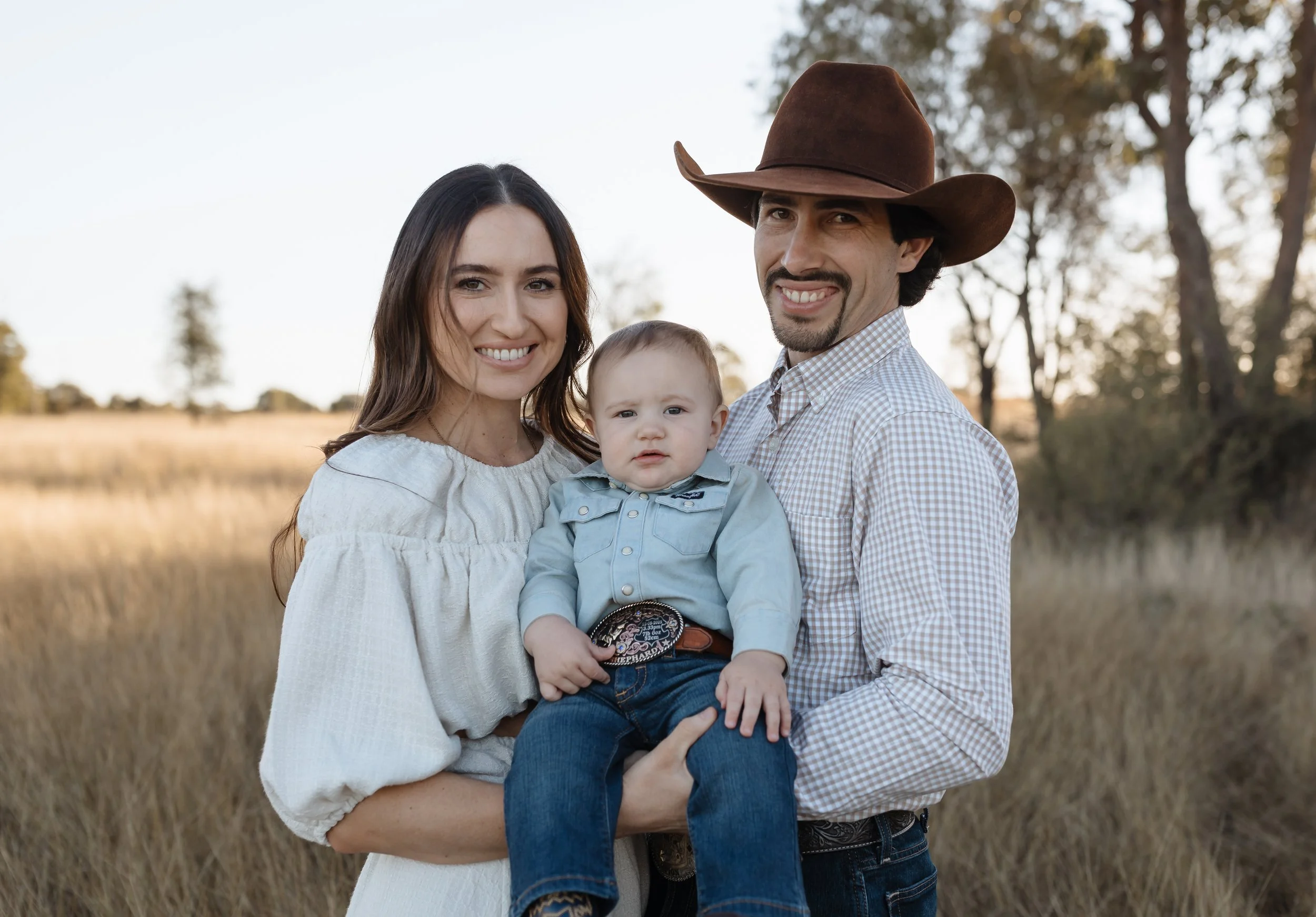 Family of three outdoors in a field with trees in the distance, woman holding a young boy, man wearing a cowboy hat.