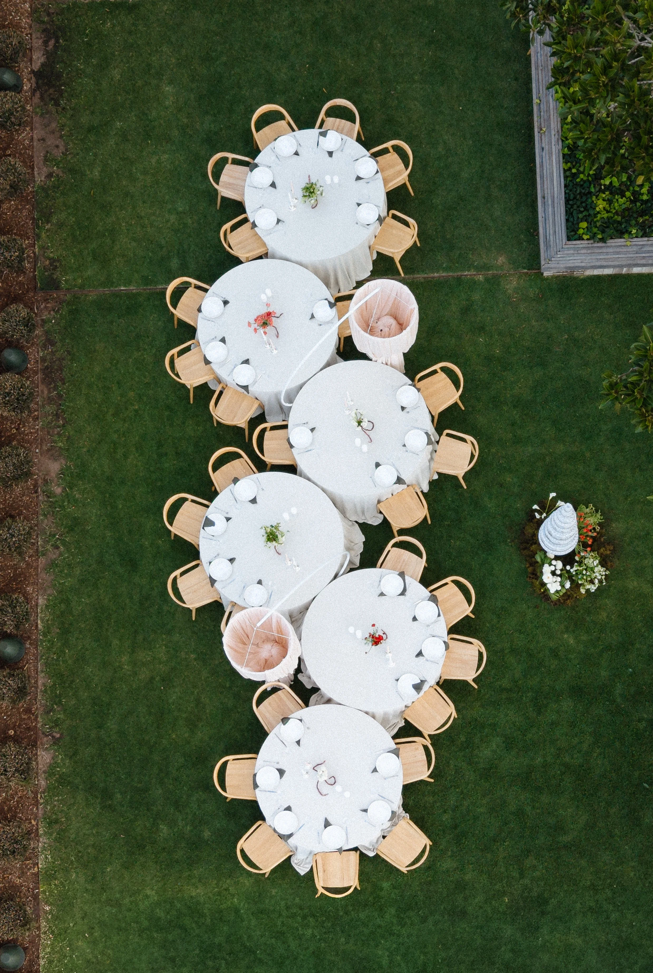 Overhead view of four round banquet tables with white tablecloths and wooden chairs, set up outdoors on a grassy area, with small flower arrangements in the center of each table.