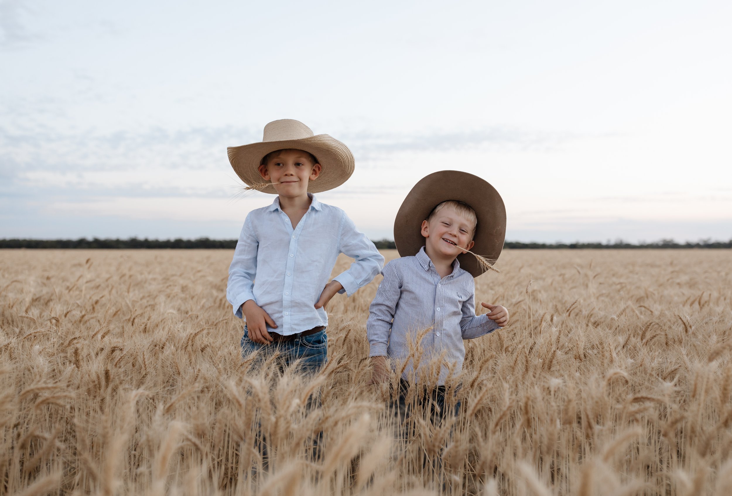 Two young boys standing in a wheat field wearing large hats, with a cloudy sky in the background.
