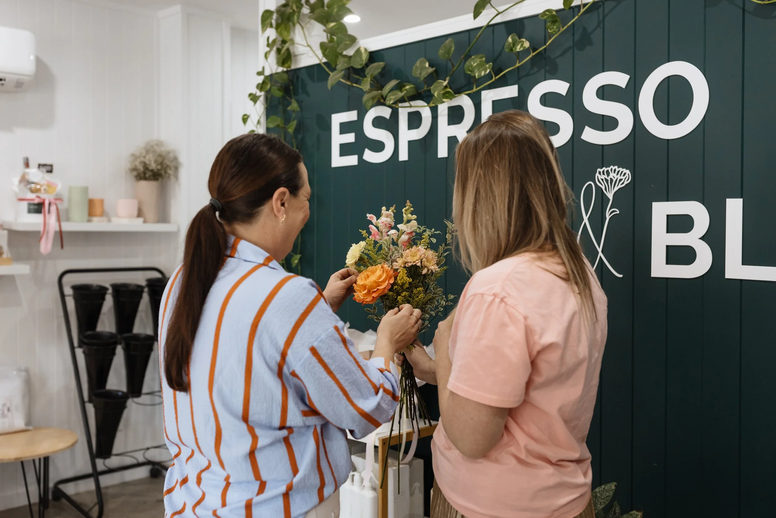 Two women exchanging flowers in a cafe with a dark green wall and large white 'ESPRESSO' sign.