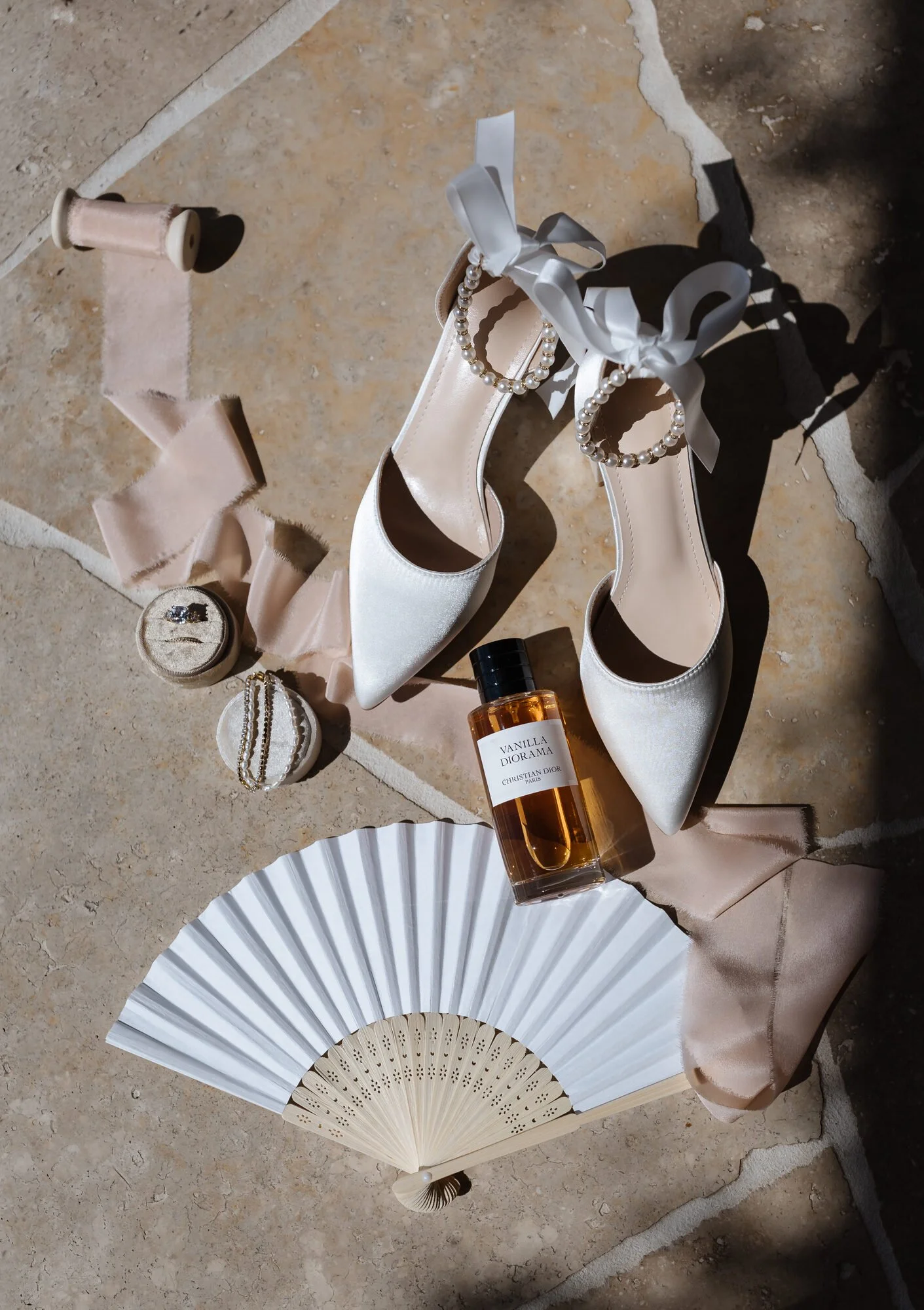 Cream-colored high-heeled shoes decorated with pearl straps and satin bows, a bottle of vanilla Diorama perfume, a pearl bracelet, a satin ribbon, a clutch with a ring, and two folding fans, all arranged on a textured stone surface with soft beige fa