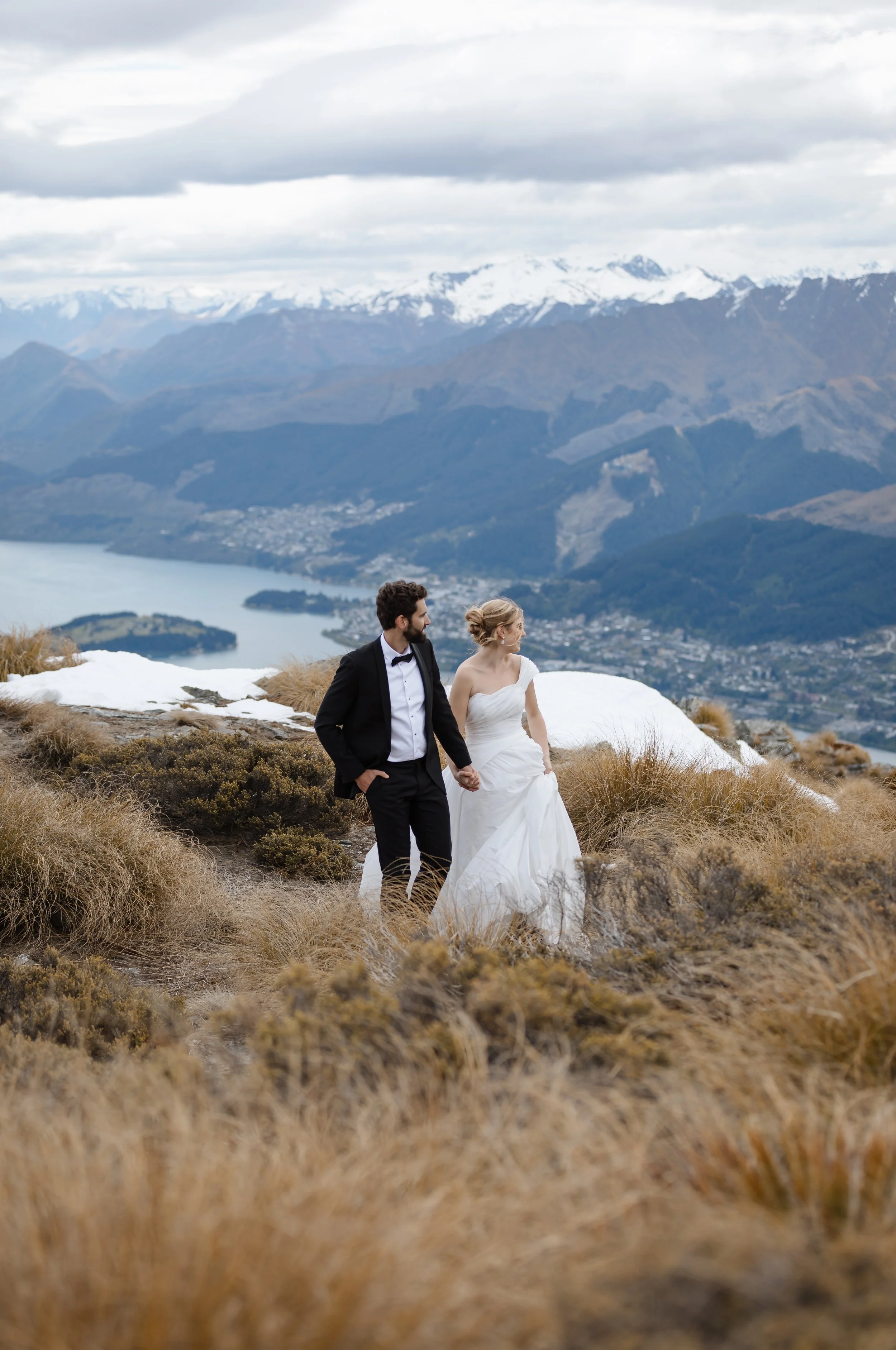 A bride and groom holding hands while walking in a mountainous landscape with snow-capped peaks and a lake in the background.