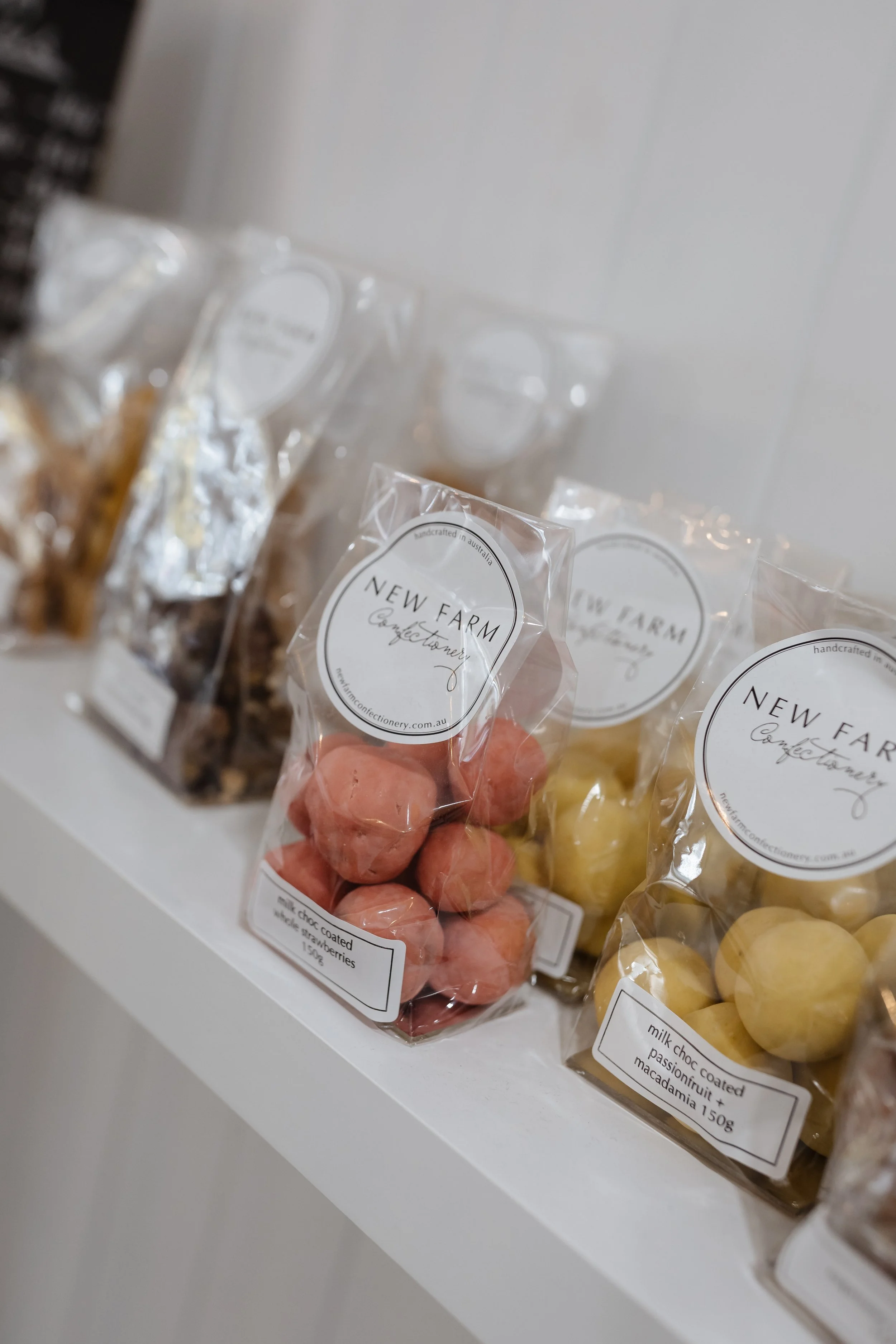 Various packages of handmade confectionery sweets and chocolates on a white shelf, including pink milk chocolate coated strawberries and yellow passionfruit macadamia, all labeled 'New Farm Confectionery'.