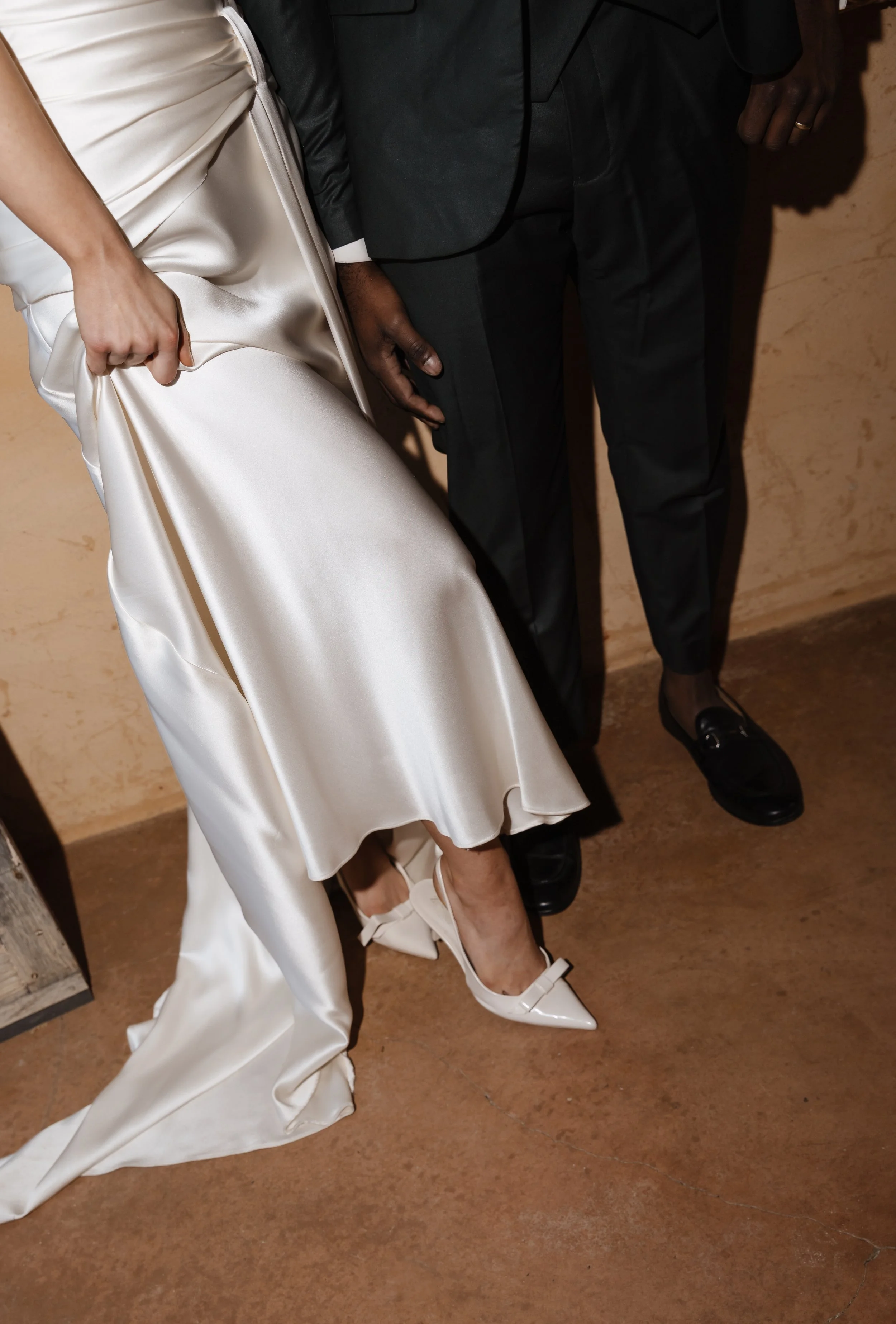 Close-up of two people dressed in formal attire, one in a white satin gown and the other in black dress shoes and suit, standing on a brown floor.