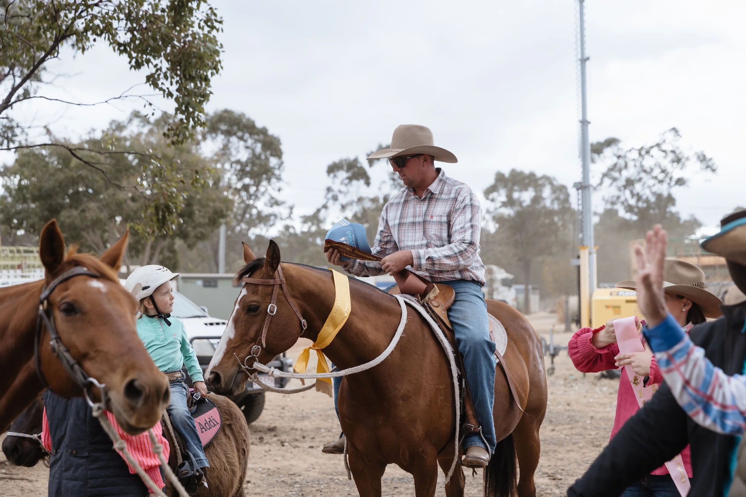 A man wearing a cowboy hat and sunglasses is riding a horse, holding a ribbon and a booklet, surrounded by children on ponies, at an outdoor event with trees and cloudy sky in the background.