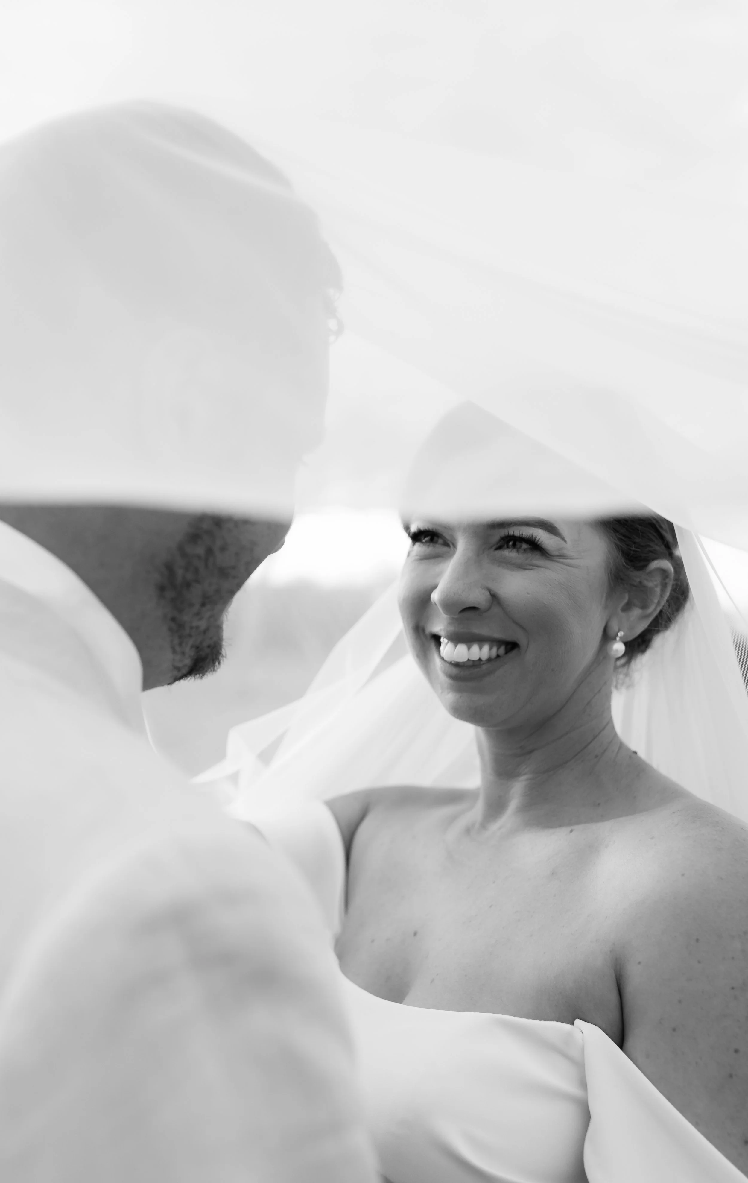 A black and white photograph of a smiling woman looking at a man, with a sheer fabric draped over both of them, highlighting an intimate moment.