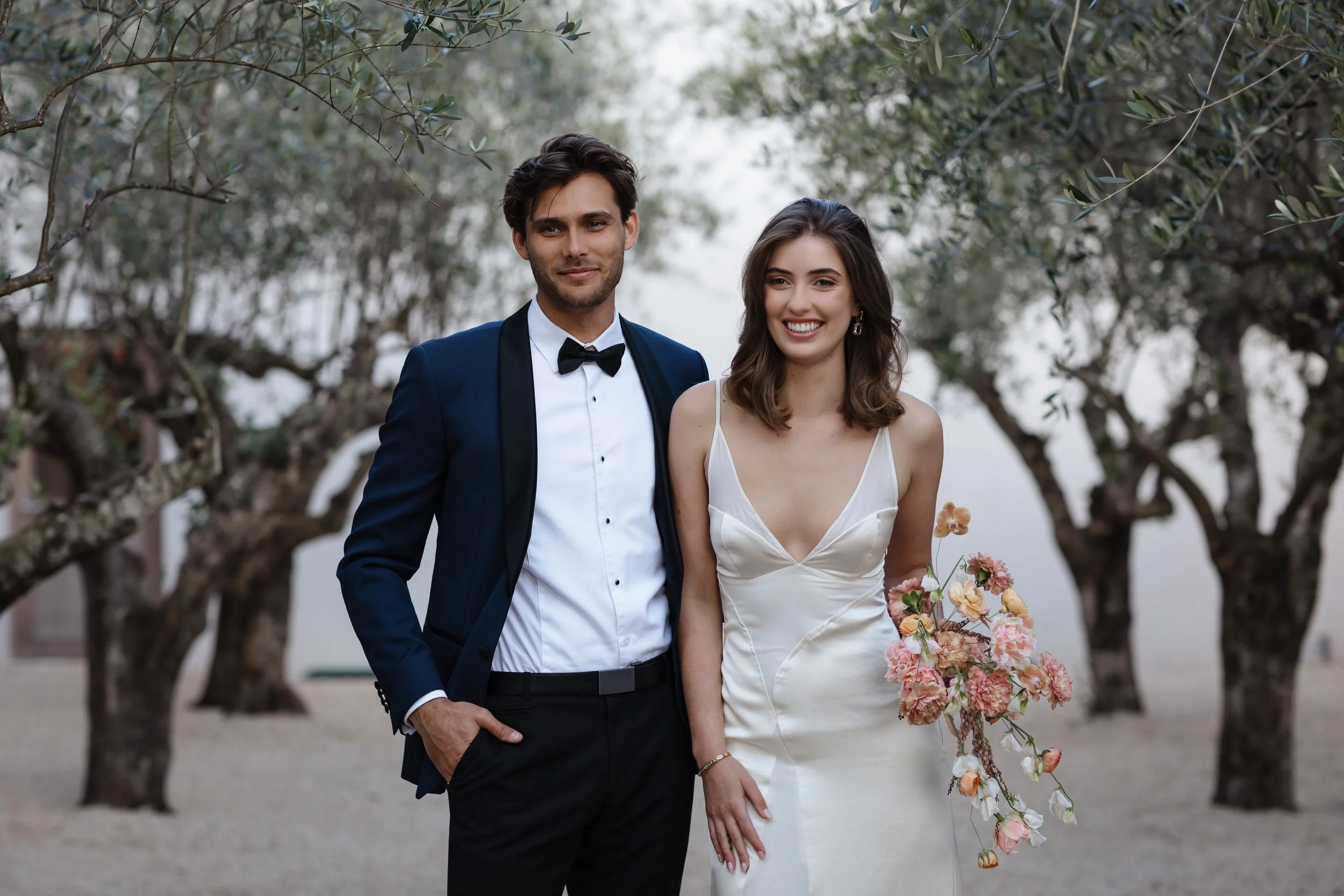 A smiling couple dressed in wedding attire walking outdoors among trees, with the man wearing a dark tuxedo and the woman in a white wedding dress holding a bouquet of pastel-colored flowers.