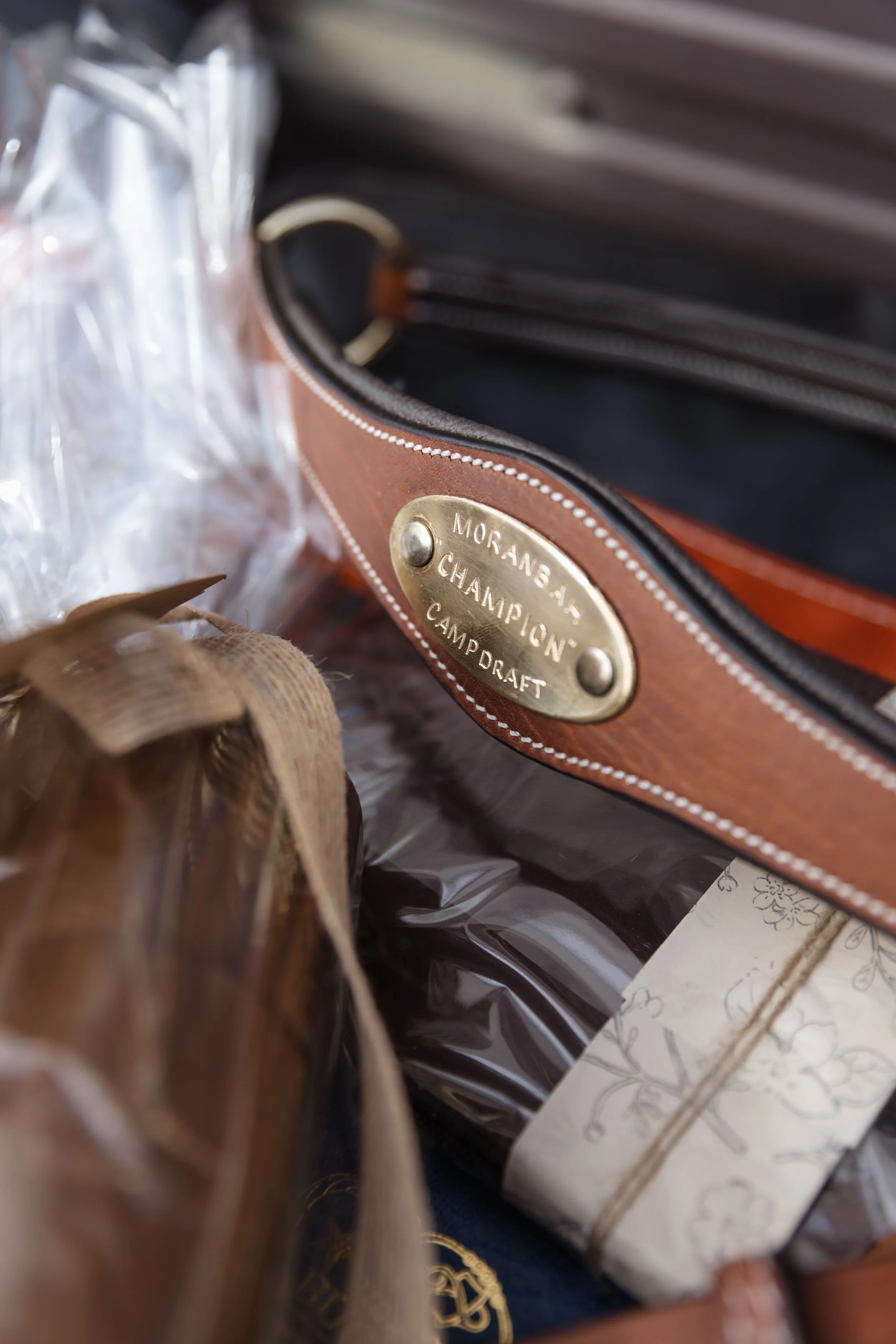 Brown leather bag with gold plaque that says 'Morane, Champ, Camp Draft' next to wrapped wrapped gift and a dried flower.