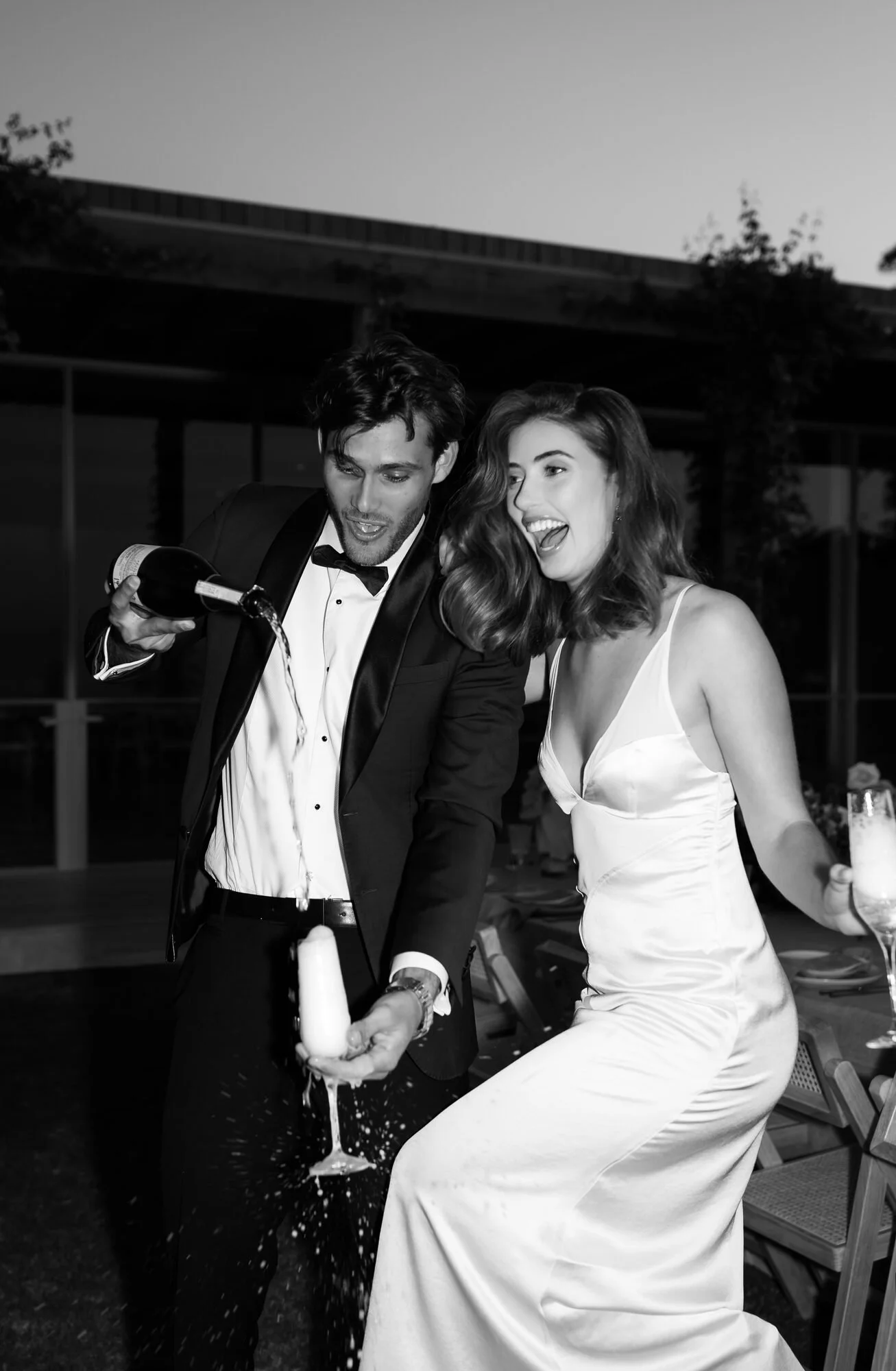 A man in a tuxedo and a woman in a white dress celebrate at a party, pouring champagne into a glass.