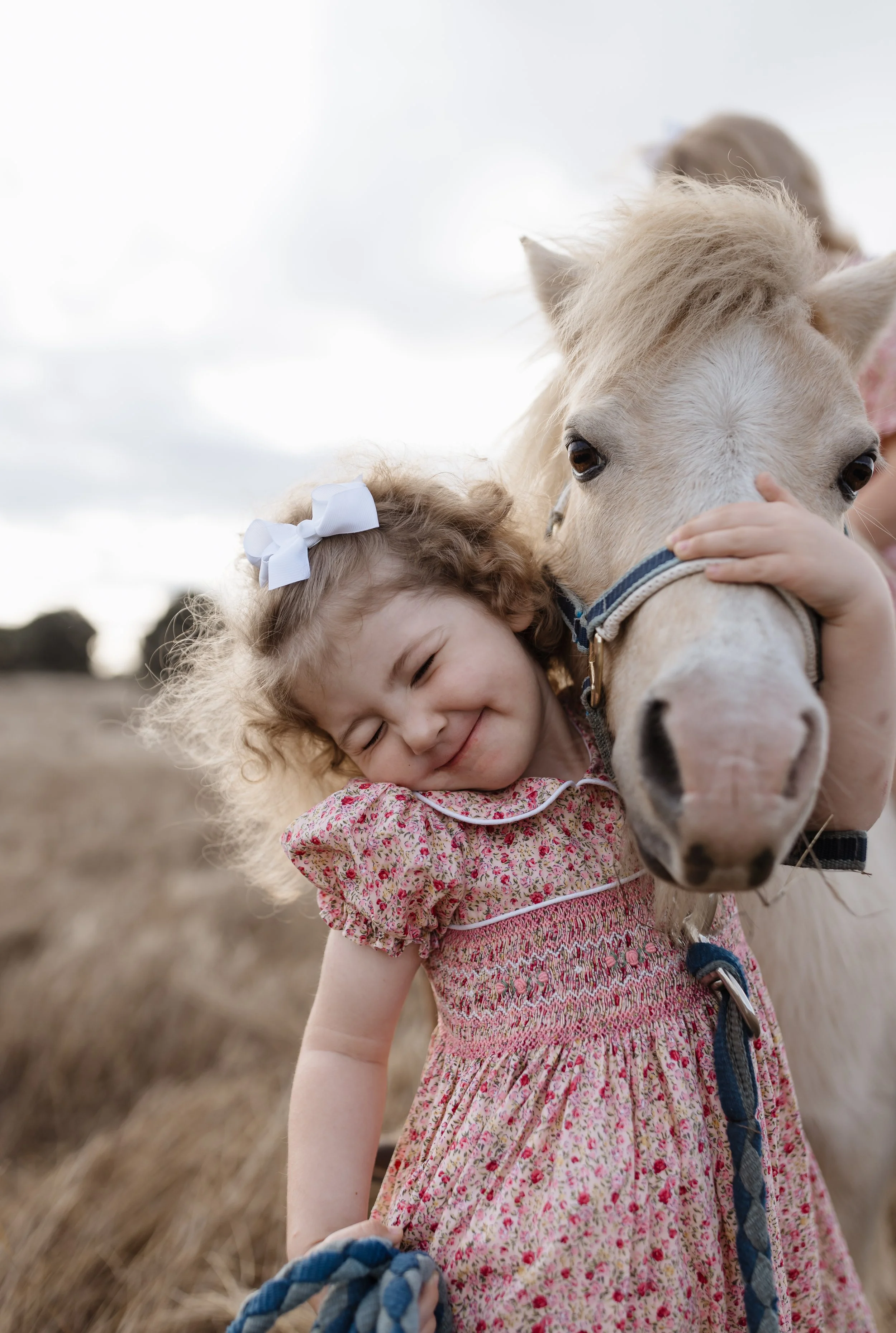 A young girl in a floral pink dress hugging a white pony in a field with tall grass and overcast sky.