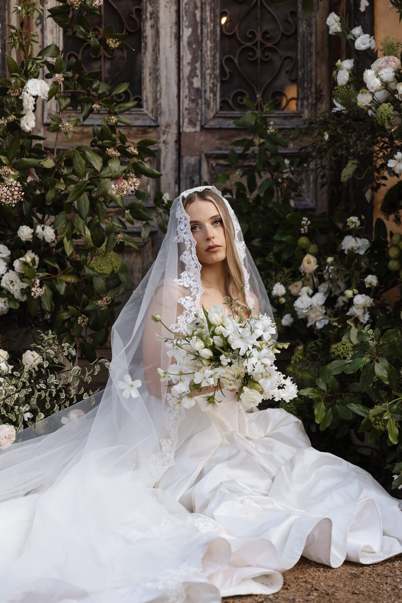 A young woman wearing a wedding dress and lace veil sits amidst white and green flowers in front of a weathered wooden door with decorative wrought iron details.