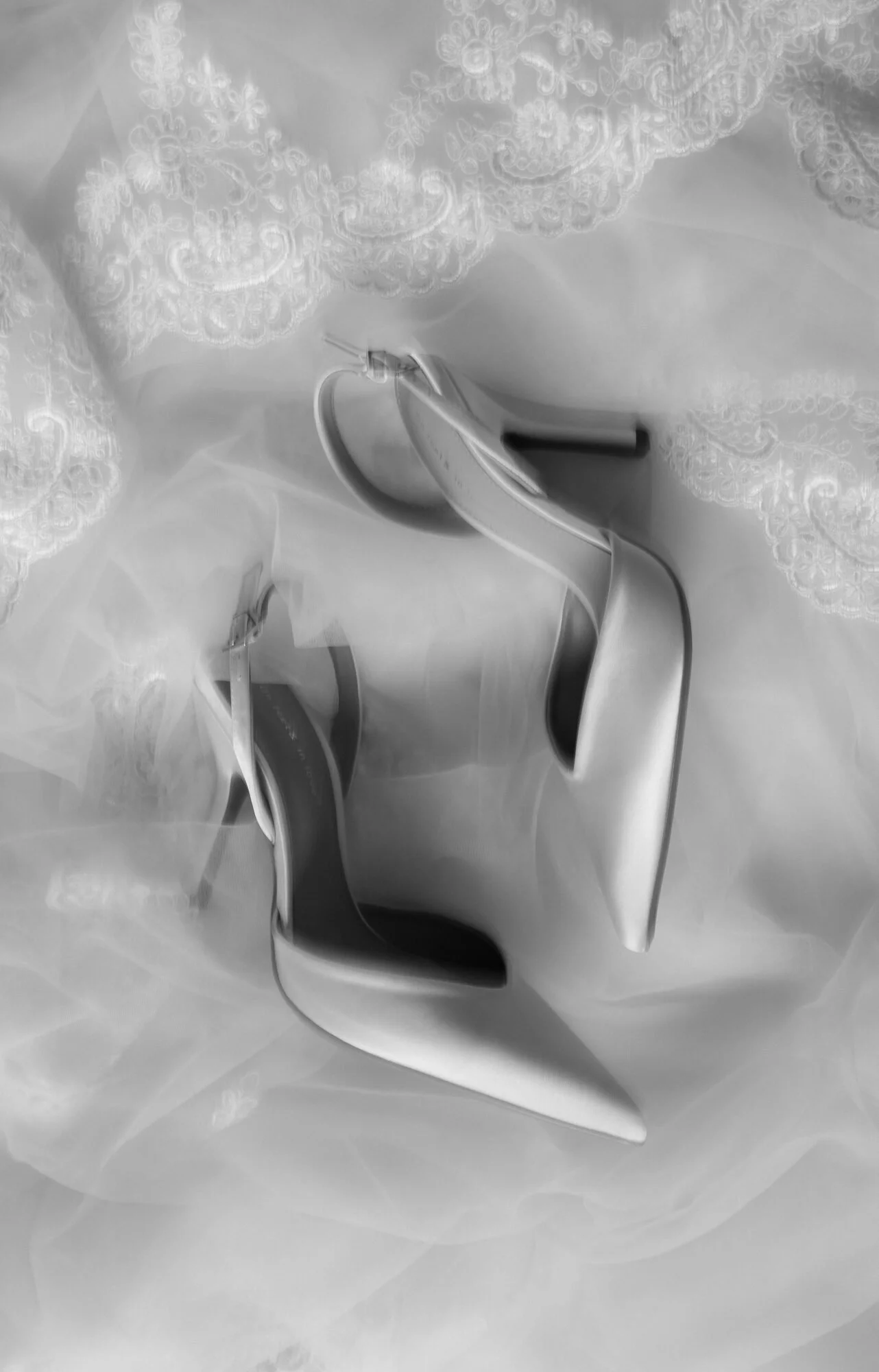 A metallic high-heeled shoe sitting on a white fabric with lace embroidery, with a thin heel and pointed toe.