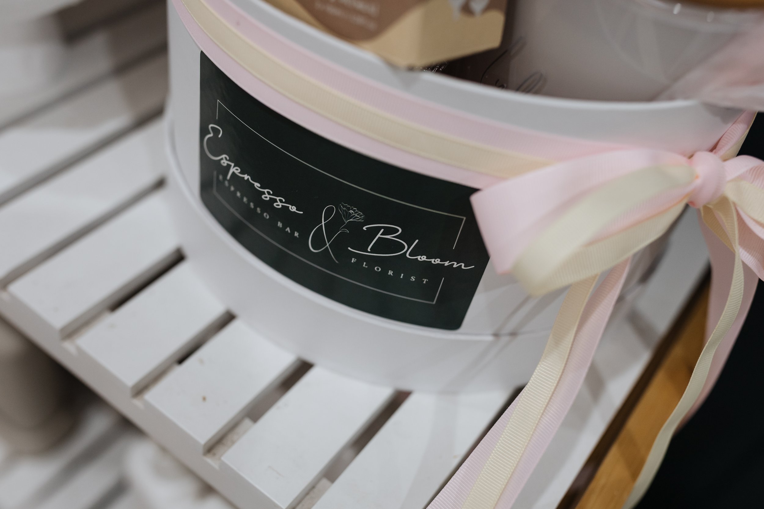 Close-up of a decorated gift box with a pink and cream ribbon, placed on a white slatted surface.