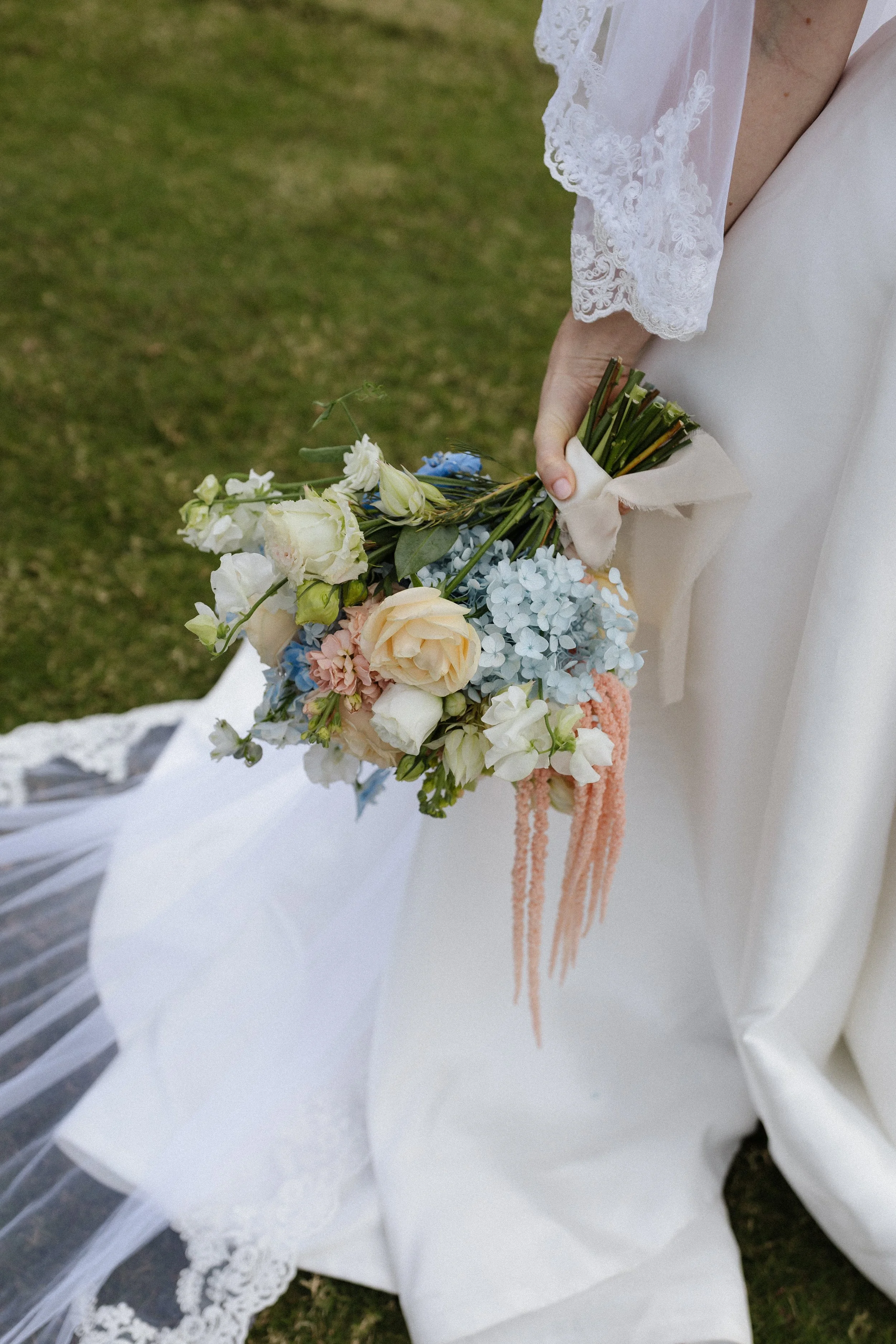 Close-up of a bride holding a pastel-colored floral bouquet, with part of her wedding dress and lace sleeve visible.