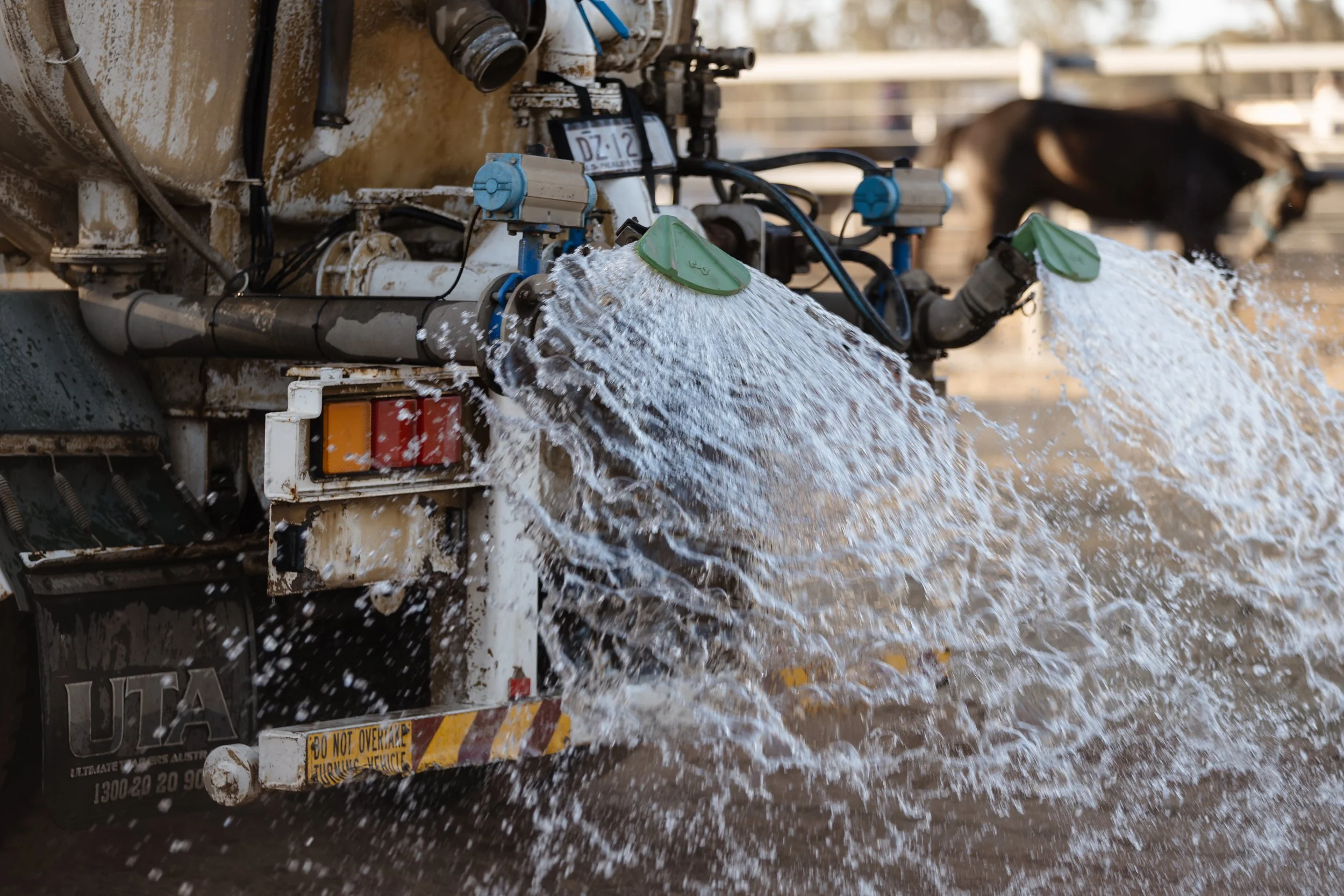A water truck spraying water onto the ground with a motorcycle blurred in the background.