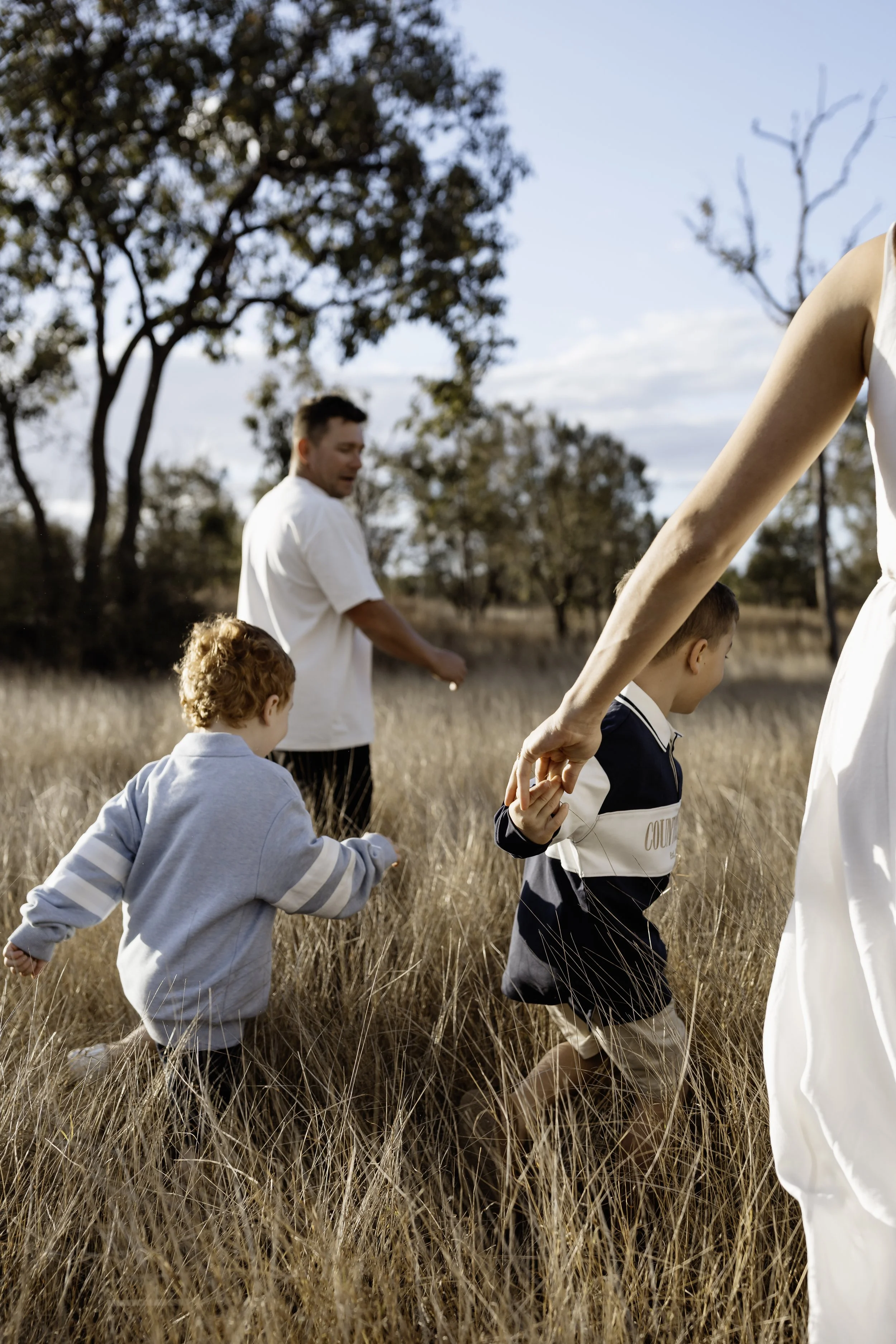 A family of four holding hands and walking through a field of tall grass in a rural area with trees and a blue sky in the background.